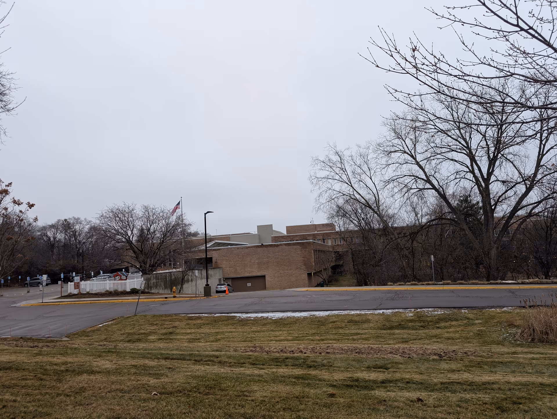 Exterior view of a large brick building with an American flag on a flagpole, surrounded by leafless trees and a grassy area in the foreground under a cloudy sky.