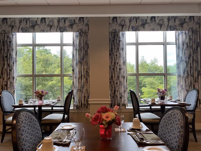 Dining room with tables set for a meal, including plates, glasses, napkins, and silverware. Each table has a small floral centerpiece in a red vase. Large windows with patterned curtains provide a view of green trees outside.