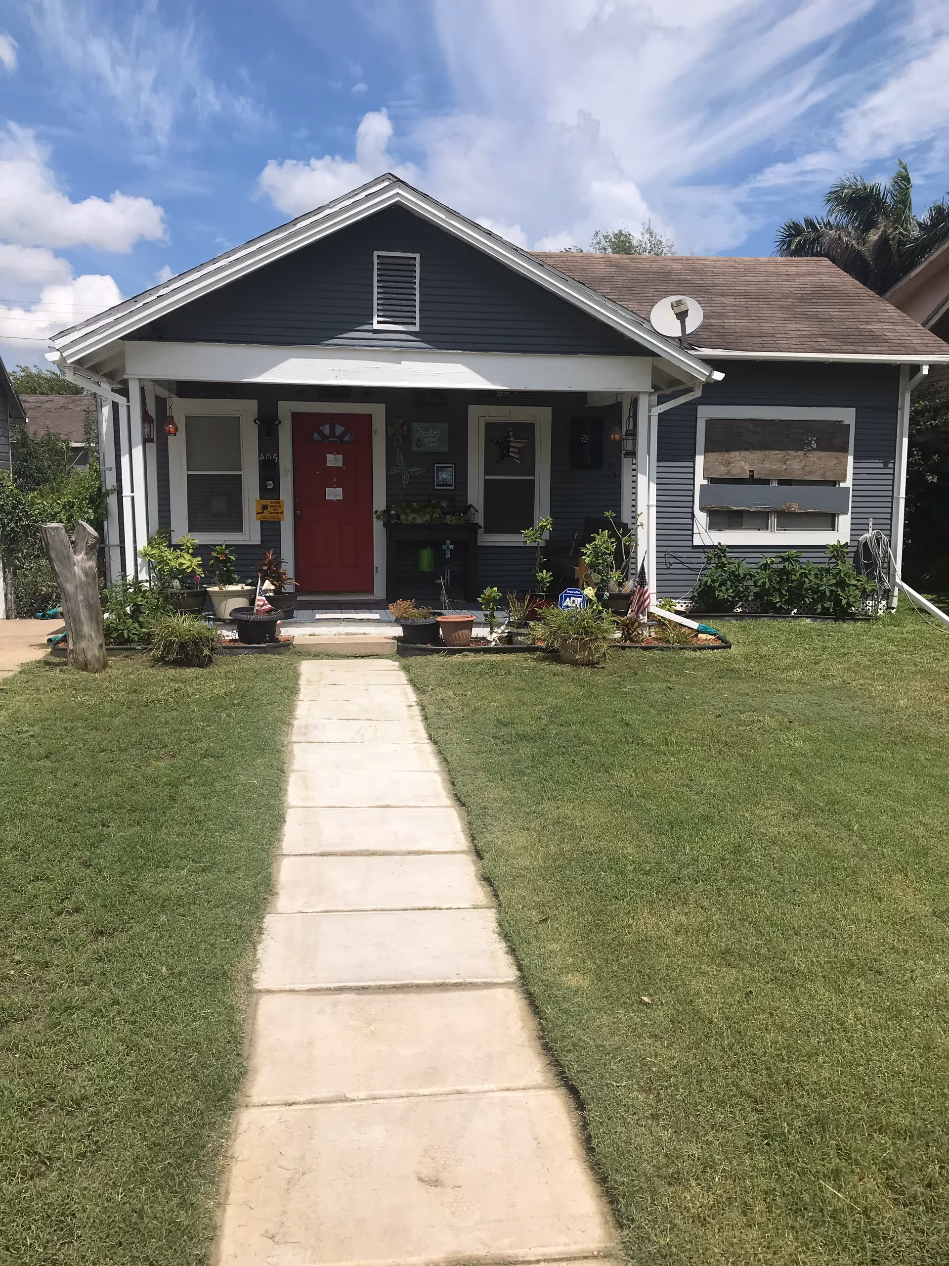 A single-story blue house with a red front door, porch plants, and a concrete walkway.