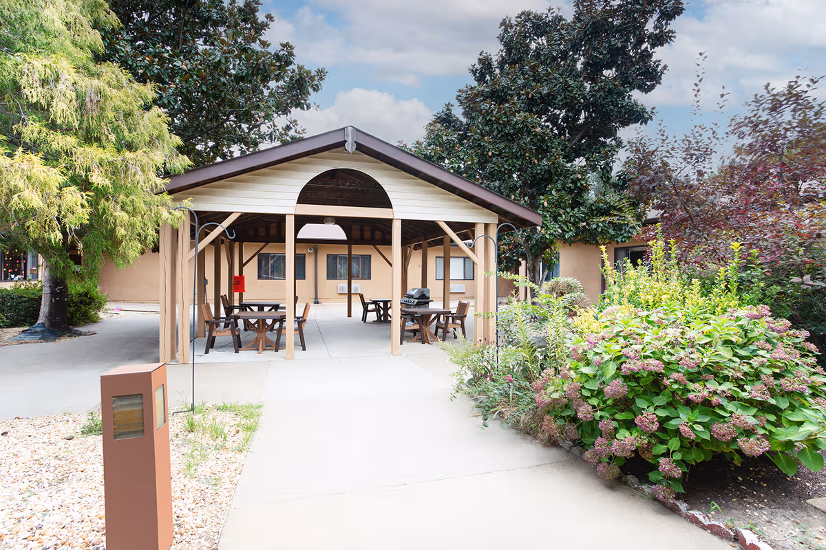 Outdoor covered pavilion with tables and chairs, surrounded by trees and flowering shrubs.