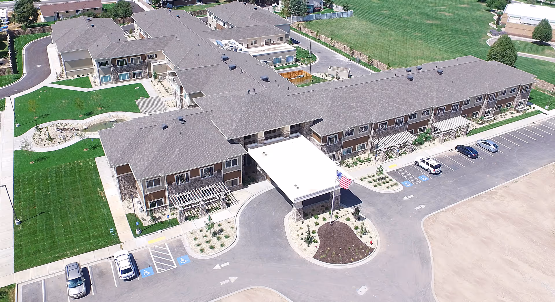Aerial view of Canyon Breeze Senior Living facility showing a large two-story building with multiple windows, a covered entrance with an American flag, surrounding landscaped green lawns, pathways, and a parking lot with several cars including handicapped parking spaces.