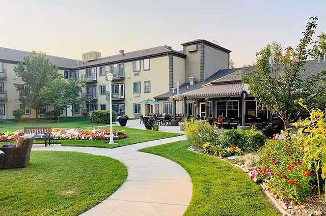 Courtyard with a curved walkway, flower beds, benches and outdoor seating in front of a multi-story senior living building.