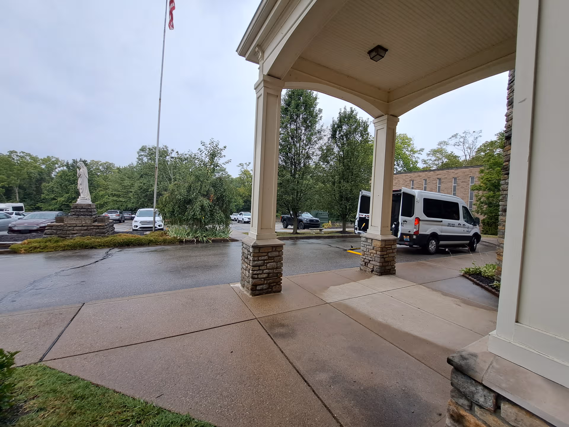 Covered entrance with stone pillars at Carmel Manor, parked vans and trees nearby.