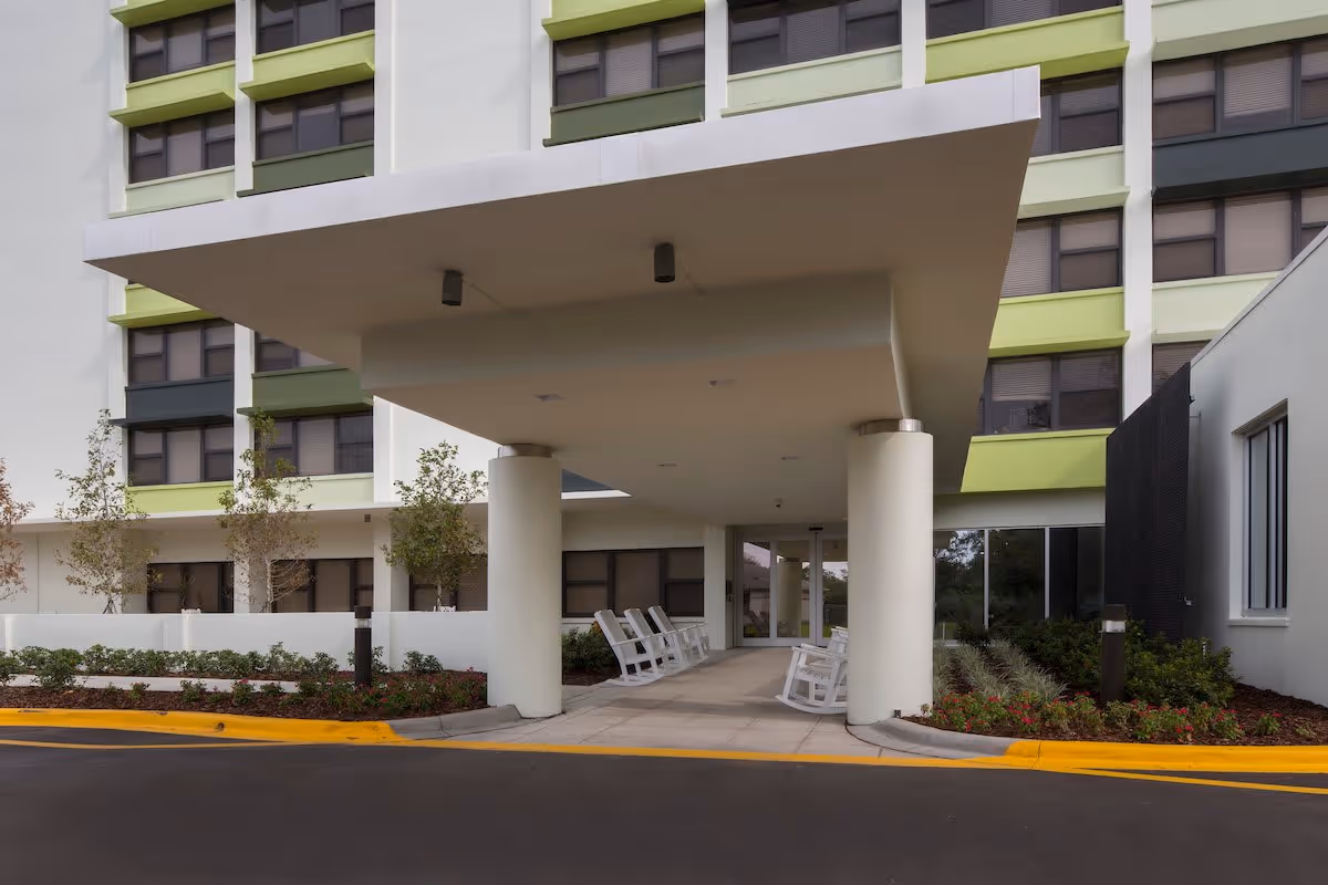 Entrance area of a senior living facility with a covered drop-off zone supported by large white columns. Several white rocking chairs are placed along the walkway under the canopy. The building exterior features multiple windows with green and white accents, and landscaped bushes and small trees surround the entrance.