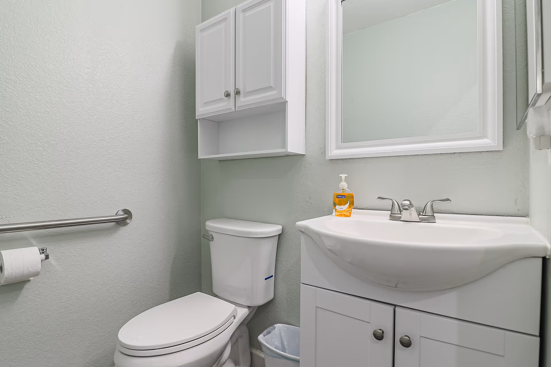 A small bathroom with a white toilet, a white vanity with a sink and faucet, a wall-mounted cabinet above the toilet, a mirror above the sink, a handrail on the left wall, and a bottle of hand soap on the sink.