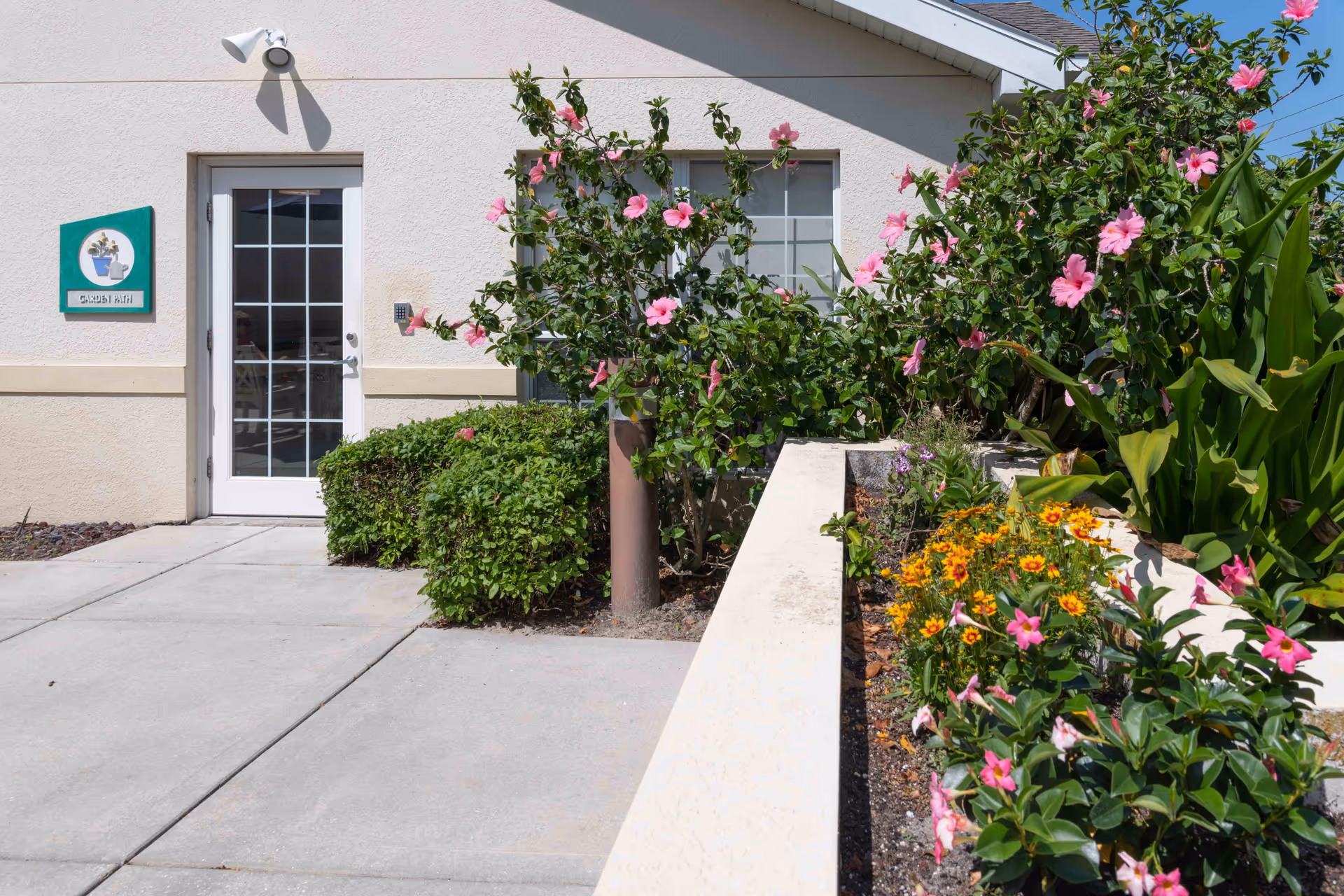 Exterior view of a building entrance with a glass door and a window. There is a green sign with a flower pot illustration and the text 'GARDEN PATH' next to the door. The area is landscaped with bushes and flowering plants, including pink and yellow flowers, along a concrete walkway.