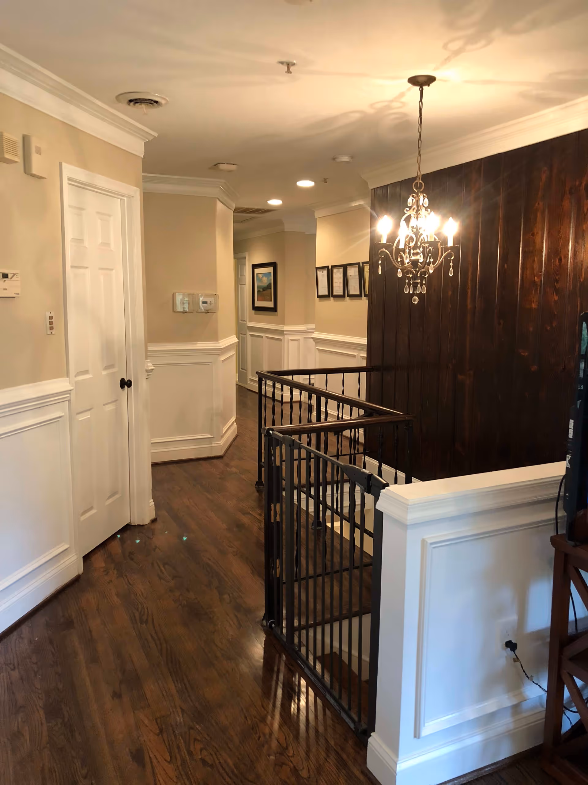 Interior hallway of a senior living facility with wooden floors, beige walls with white wainscoting, a white door on the left, framed pictures on the walls, a black metal safety gate at the top of a staircase, and a decorative chandelier hanging from the ceiling.