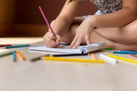 Close-up of hands drawing on paper with colorful pencils on a table indoors.