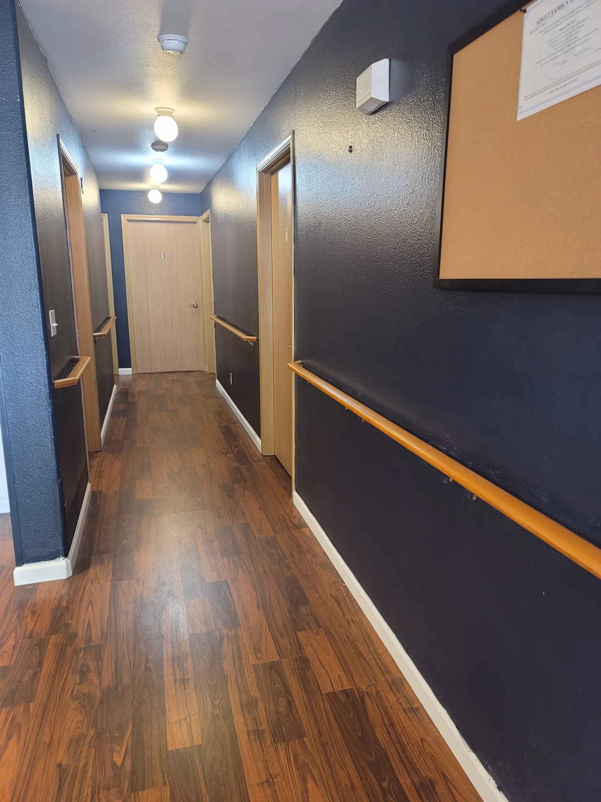 Narrow hallway in a senior living facility with dark blue walls, wood floors, and handrails.
