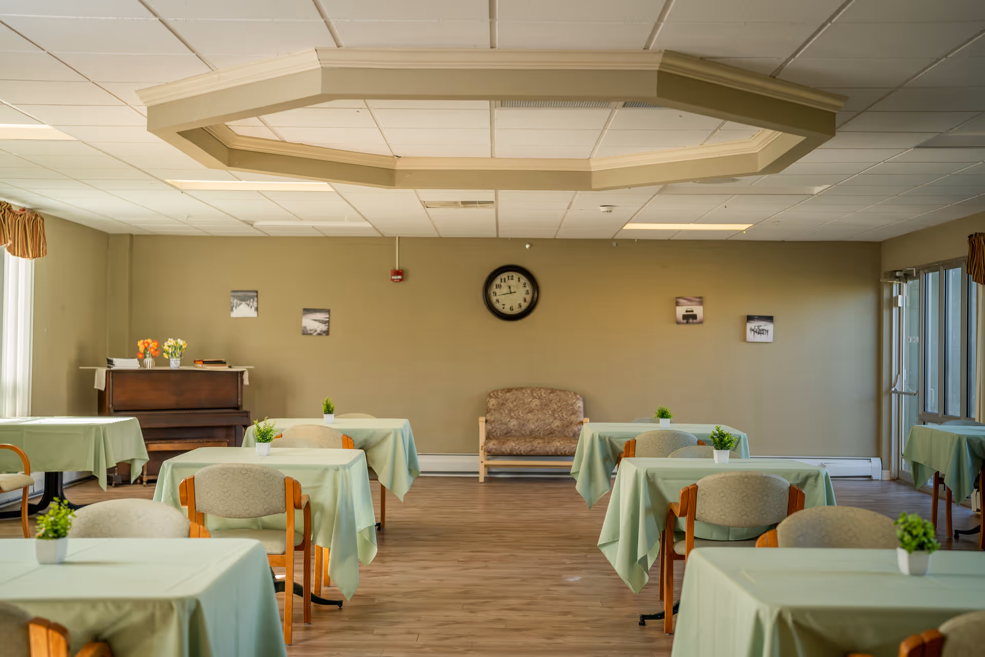 Senior living dining room with green tablecloths, chairs, piano, couch, and clock.