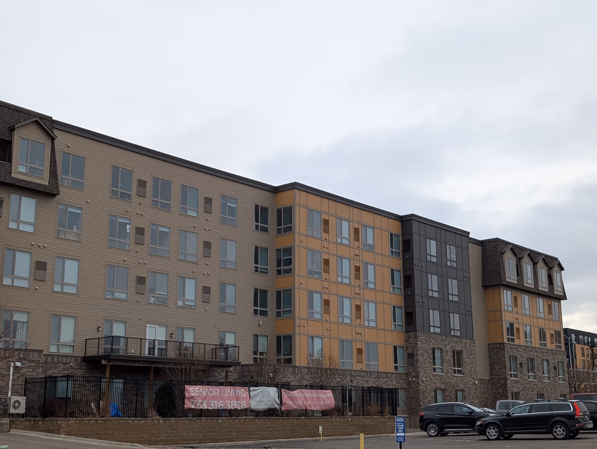 Exterior view of a multi-story senior living facility building with beige, orange, and dark gray panels. Several windows are visible, along with a fenced area and parked cars in front. Banners on the fence display contact information and the name Global Pointe Senior Living.