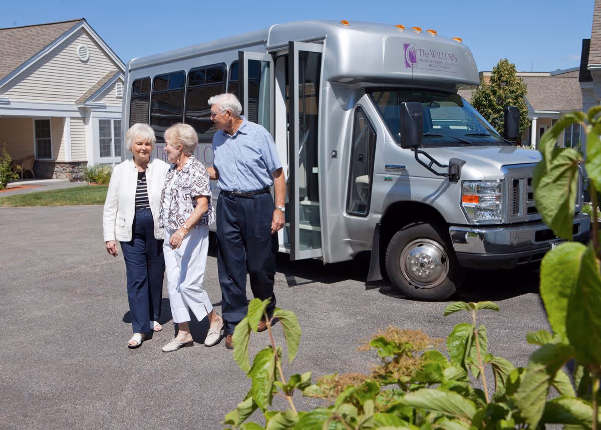 Three elderly people, two women and one man, walking together outside near a silver shuttle bus with The Willows Retirement Community logo on it. They are in a paved area with residential-style buildings in the background and some green plants in the foreground.