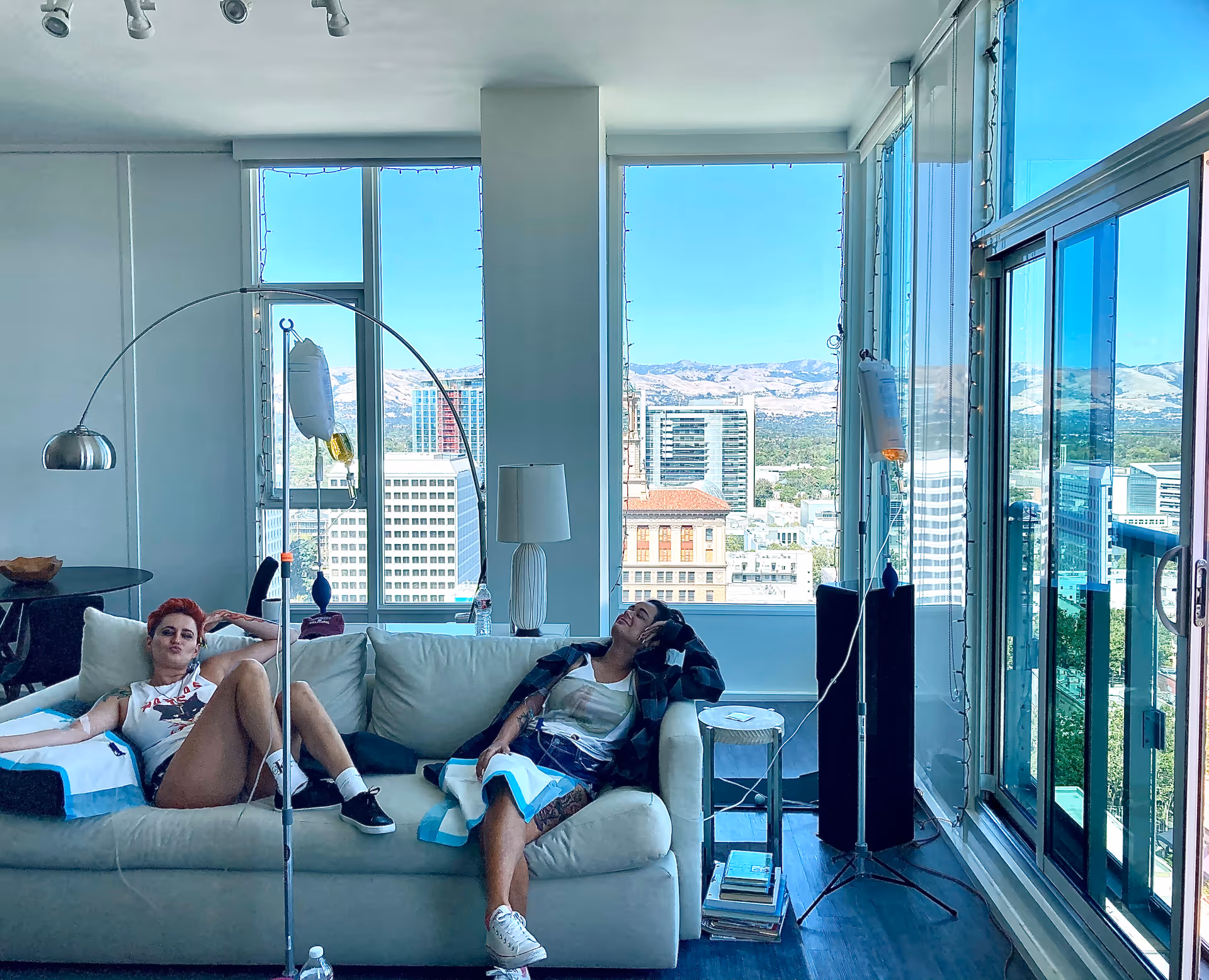 Two residents lounge on a light sofa in a bright high-rise living room with city view.