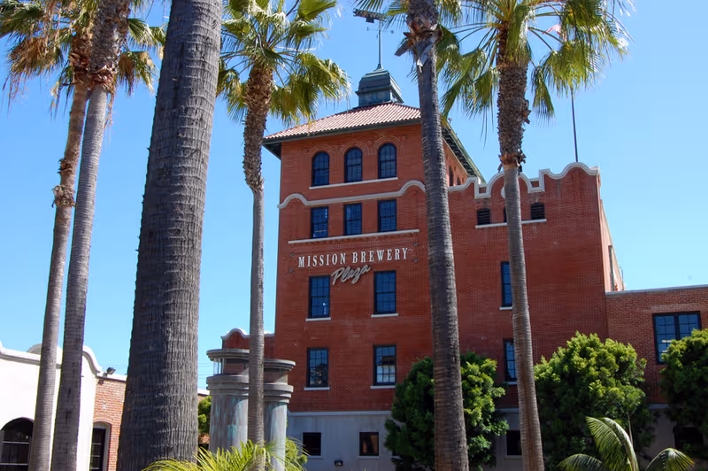 Red brick Mission Brewery building with tall palm trees in the foreground.