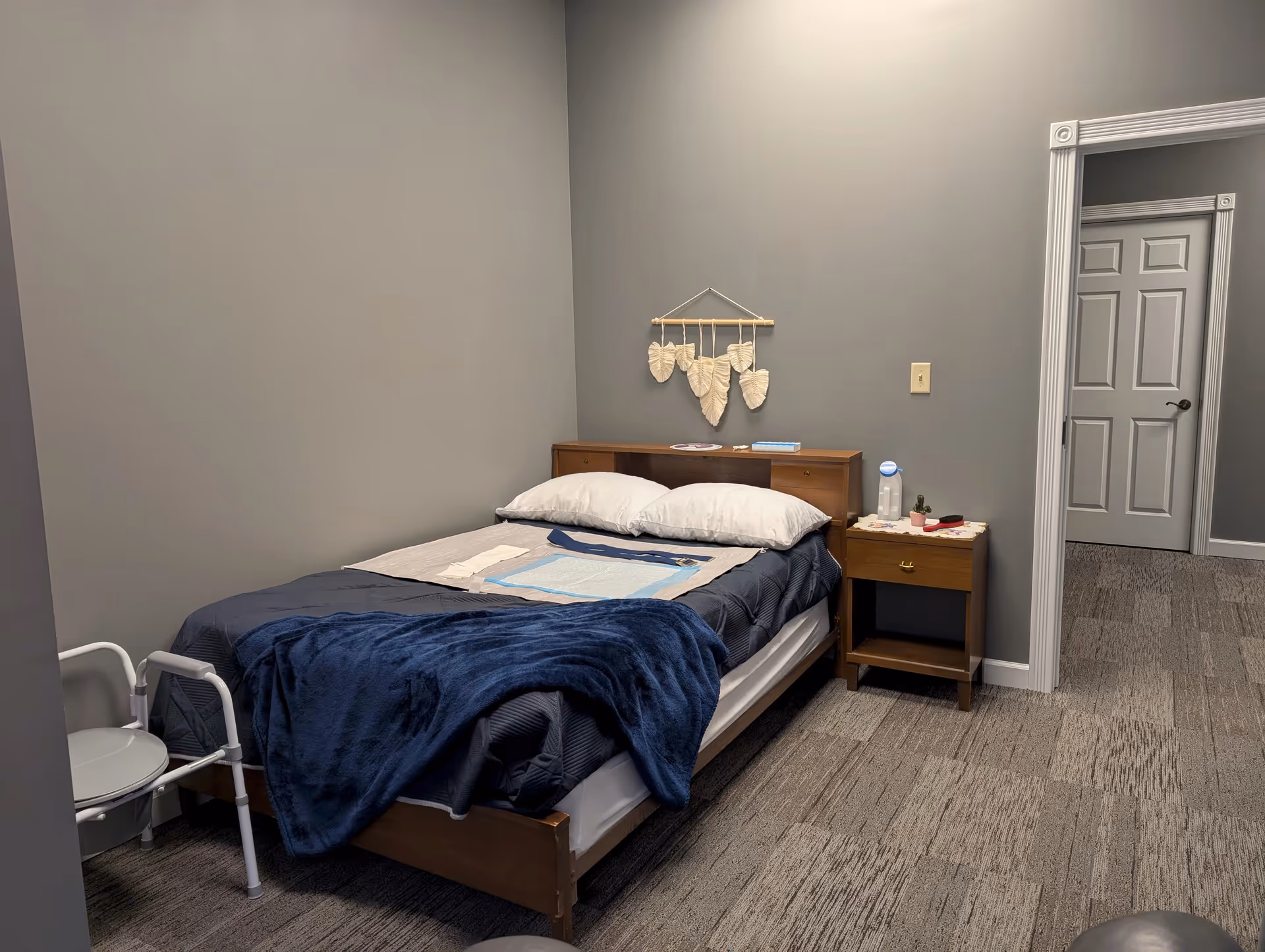 A simple bedroom in a senior facility featuring a bed, nightstand, and doorway.