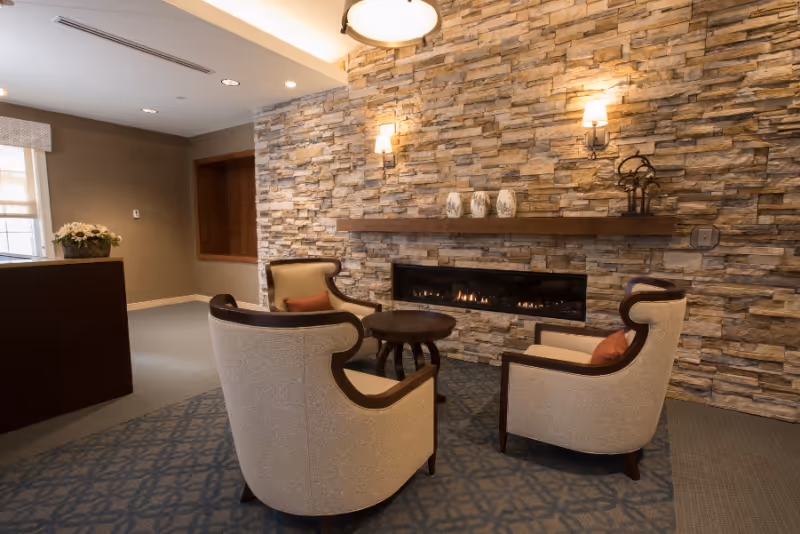 A cozy seating area with three beige armchairs arranged around a small round wooden table in front of a modern rectangular fireplace set into a stone accent wall. Above the fireplace is a wooden mantel with decorative vases and a small sculpture. The room has warm lighting from wall sconces and a ceiling light fixture, with a patterned carpet and neutral-colored walls.