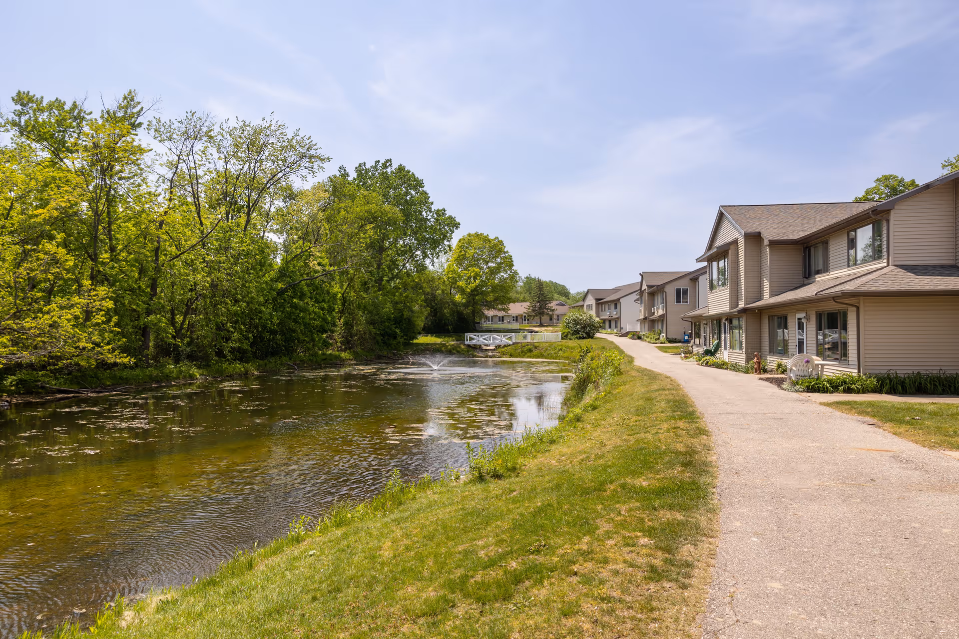 A paved pathway runs beside a pond with trees on the left and multi-unit senior living buildings on the right under a blue sky.