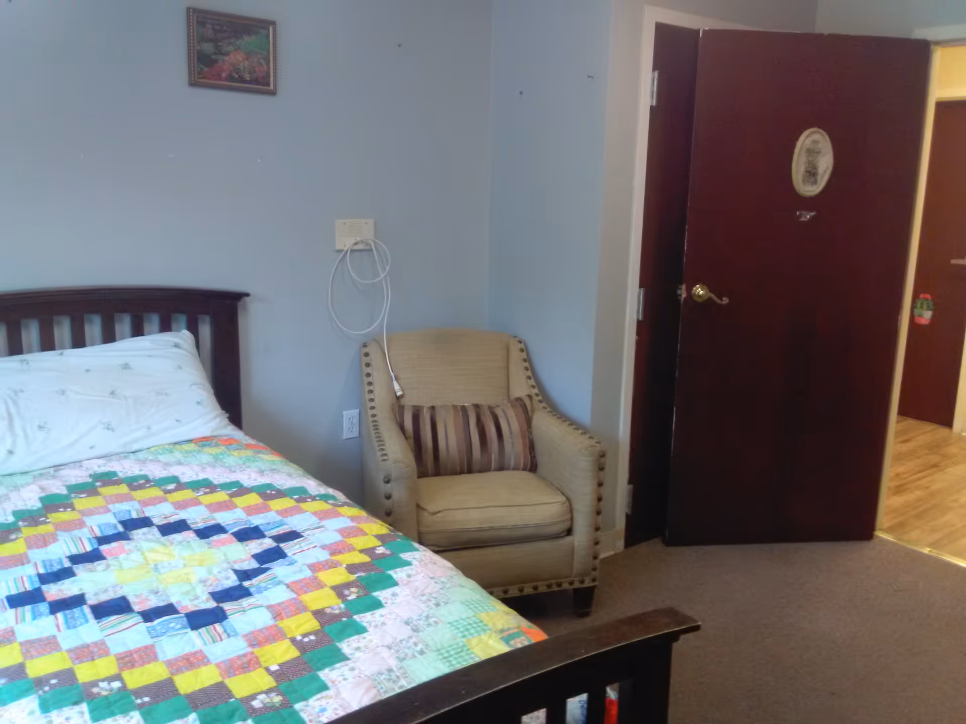 A small bedroom with a wooden bed frame and a colorful patchwork quilt on the bed. Next to the bed is a beige upholstered armchair with a striped pillow. The walls are light blue with a small framed picture hanging above the chair. A dark wooden door is open, showing a glimpse of a hallway with wooden flooring.