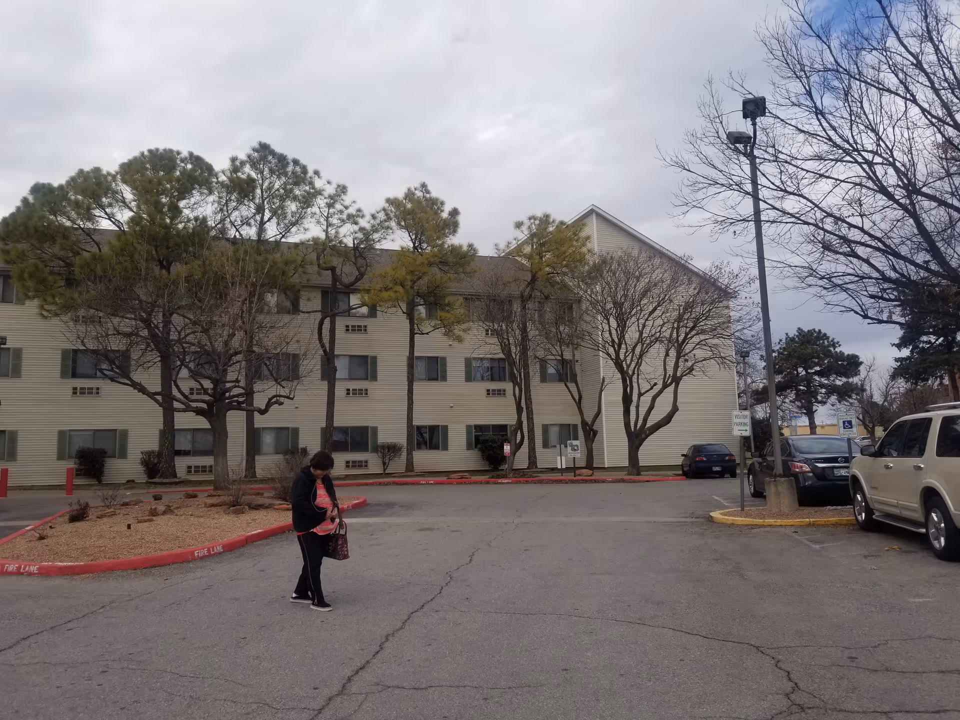 Three-story beige senior living building with trees, a parking lot, and a person walking nearby.