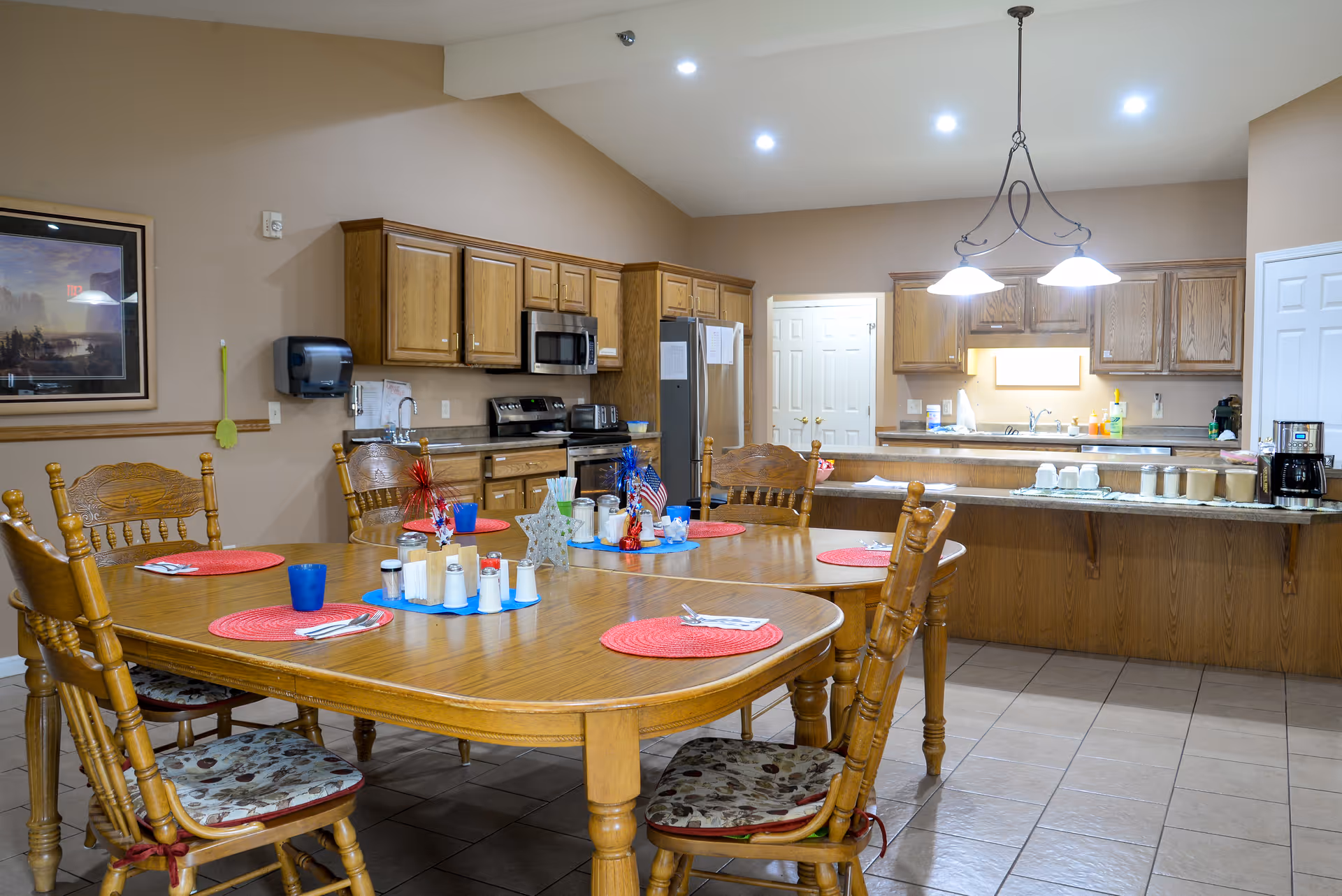 A spacious kitchen and dining area with wooden cabinets and a large wooden dining table set with red placemats, utensils, and condiment containers. The kitchen features modern appliances including a microwave, stove, and refrigerator. Overhead lights illuminate the room, and there is a coffee maker on the counter.