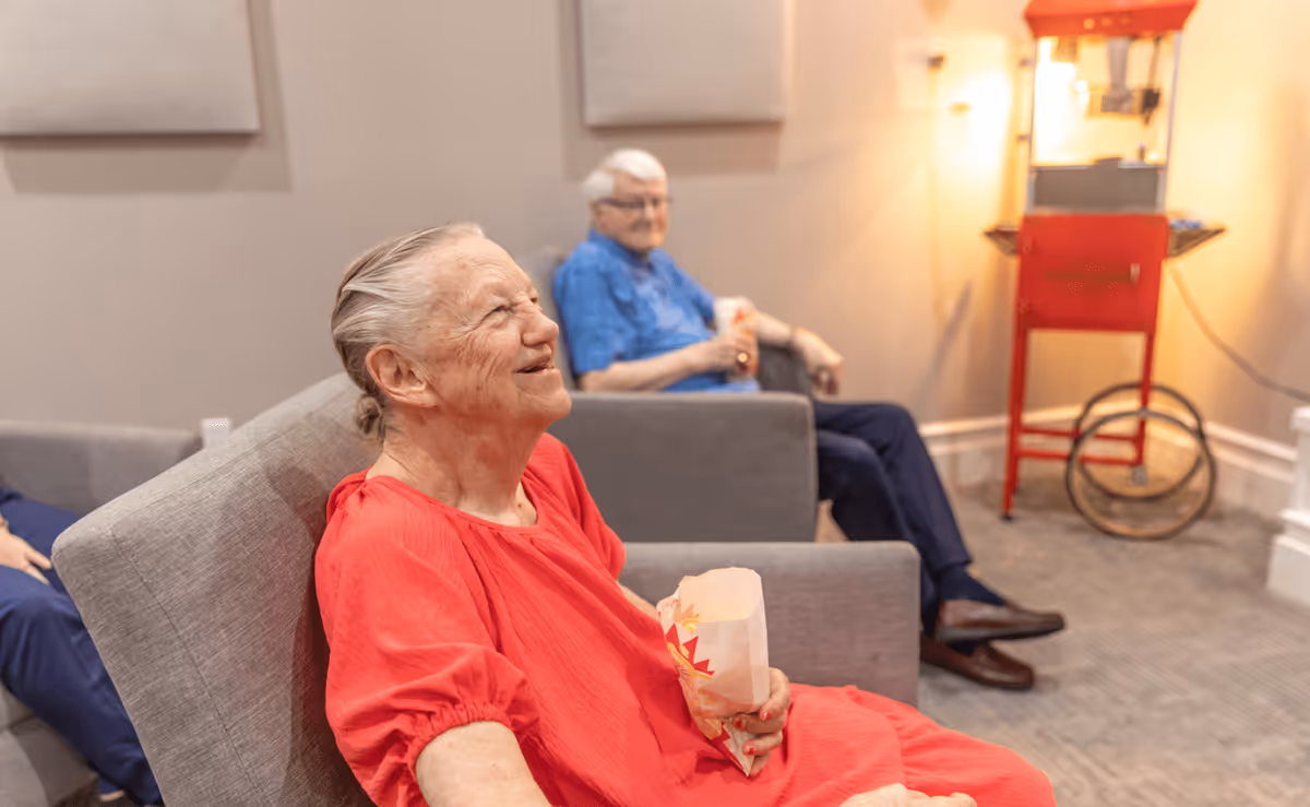 Two elderly individuals sitting comfortably in a lounge area, each holding a bag of popcorn. One person in a red shirt is smiling and looking to the side, while the other person in a blue shirt is seated further back. A red popcorn machine is visible in the background.