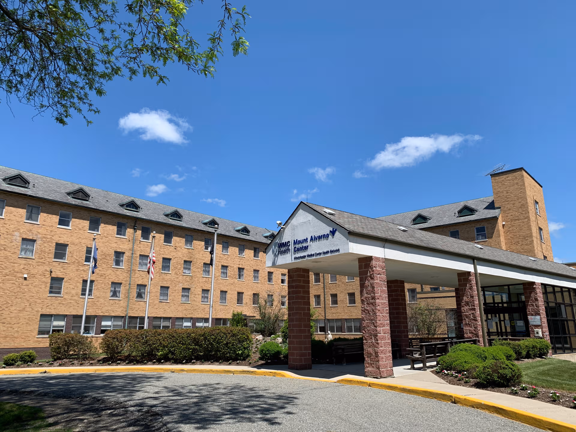 Exterior of Mt Alverno Center, brick building with a covered entrance and flags against blue sky.