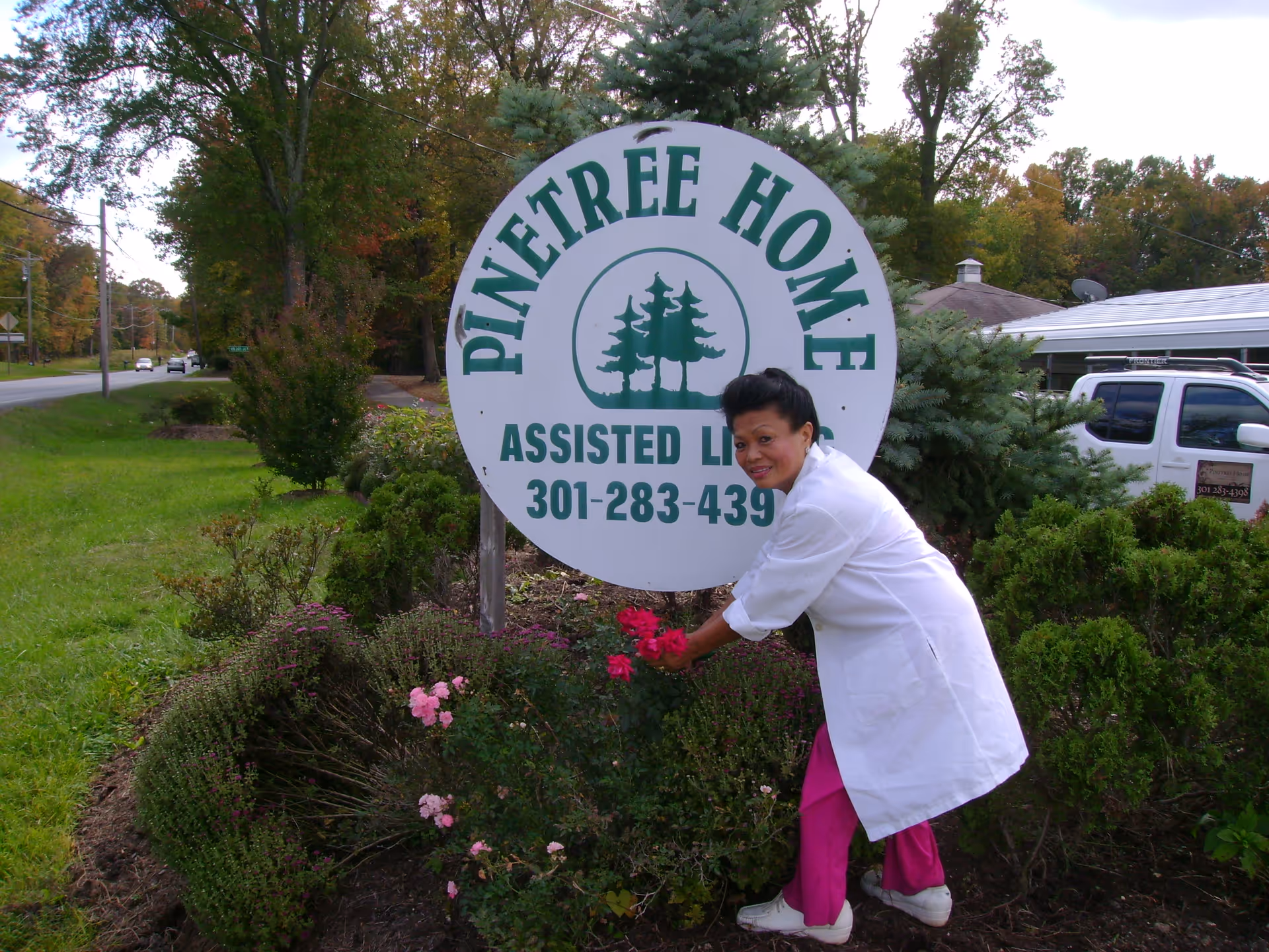 A woman in a white coat and pink pants is tending to flowers in front of a large round sign that reads 'Pinetree Home Assisted Living 301-283-439'. The sign is surrounded by bushes and trees, with a road and vehicles visible in the background.