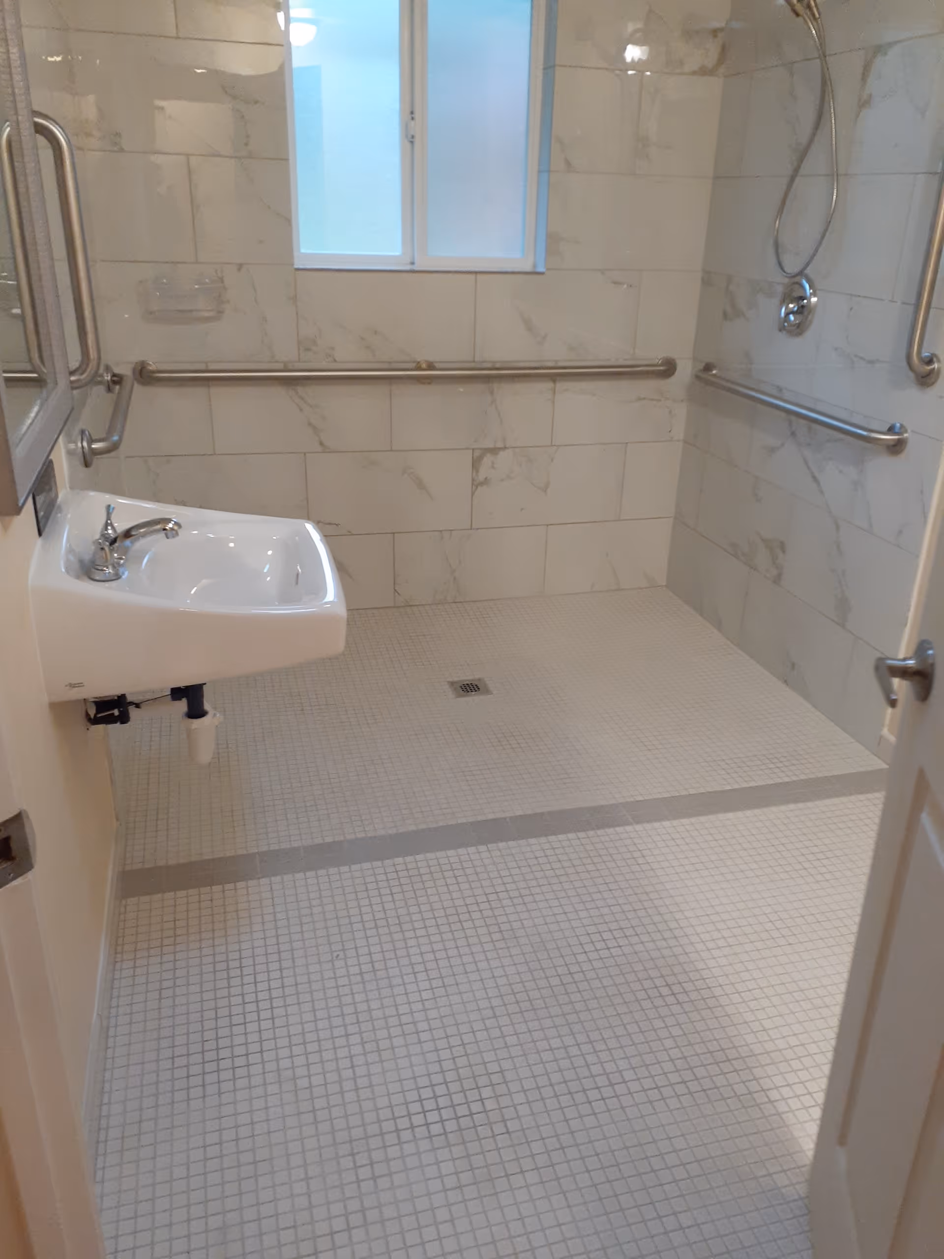 A bathroom with a walk-in shower area featuring white tiled walls with a marble pattern and small square floor tiles. There are multiple stainless steel grab bars installed on the walls for support. A small white wall-mounted sink with a faucet is visible on the left side. A frosted window is present on the back wall, allowing natural light into the room.
