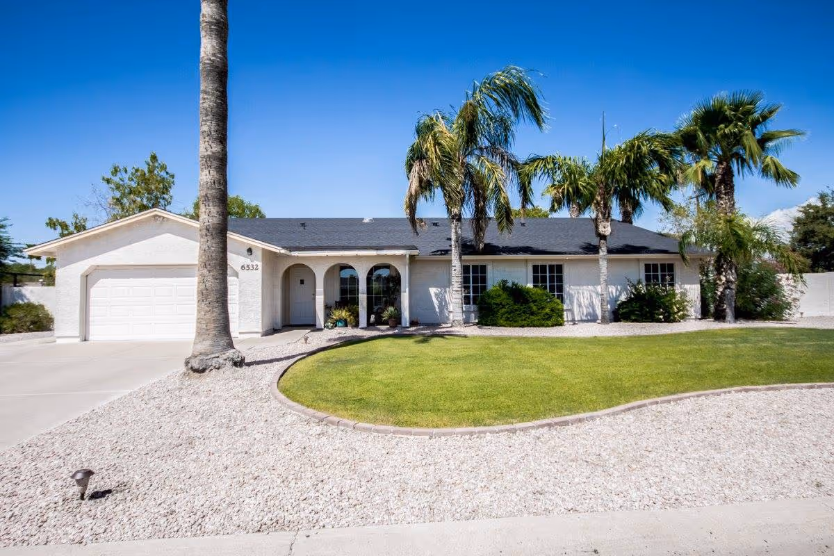 Single-story white house with a dark roof, a two-car garage, and a front yard featuring green grass, palm trees, and gravel landscaping under a clear blue sky.