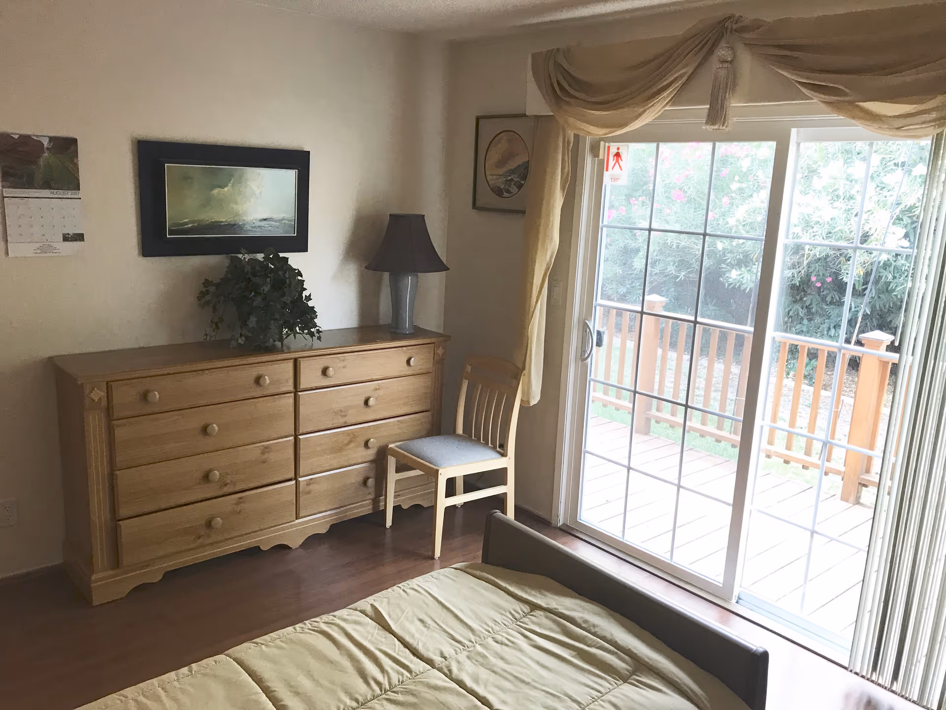 A bedroom with a wooden dresser topped with a lamp and a plant, a wooden chair next to a sliding glass door with beige curtains, and a bed with a light green comforter. Outside the door, there is a wooden deck and greenery.