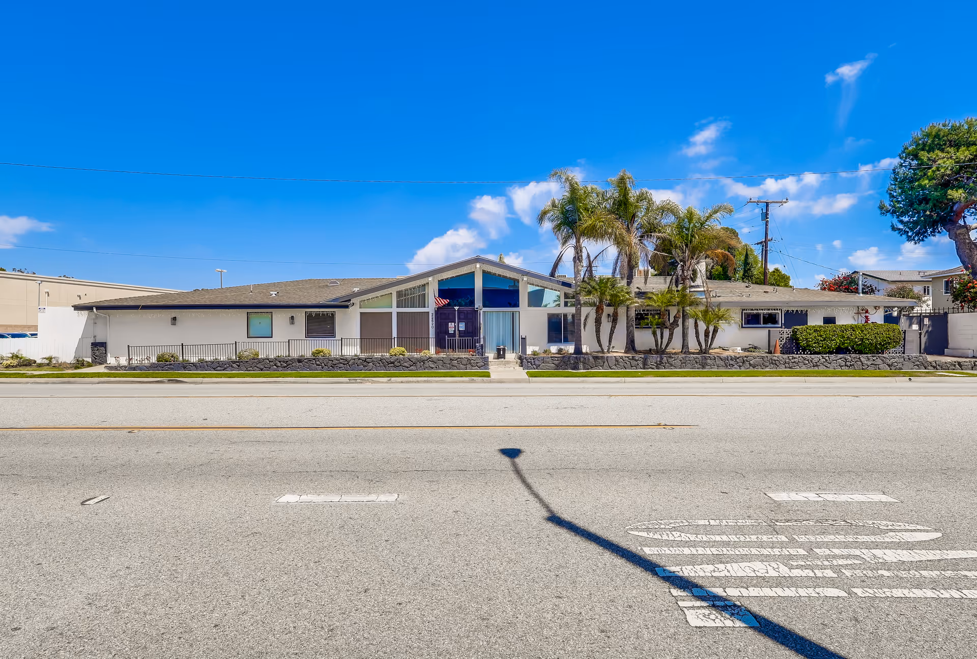 Exterior front view of a single-story building with a sloped roof, large windows, and palm trees in front. The building is situated along a street with a clear blue sky overhead.
