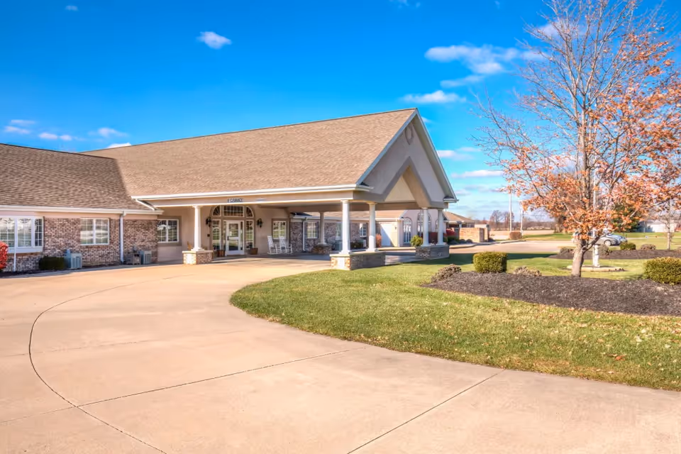 Front entrance of a single-story senior living facility with a covered porte-cochere, curved driveway, and landscaped lawn.