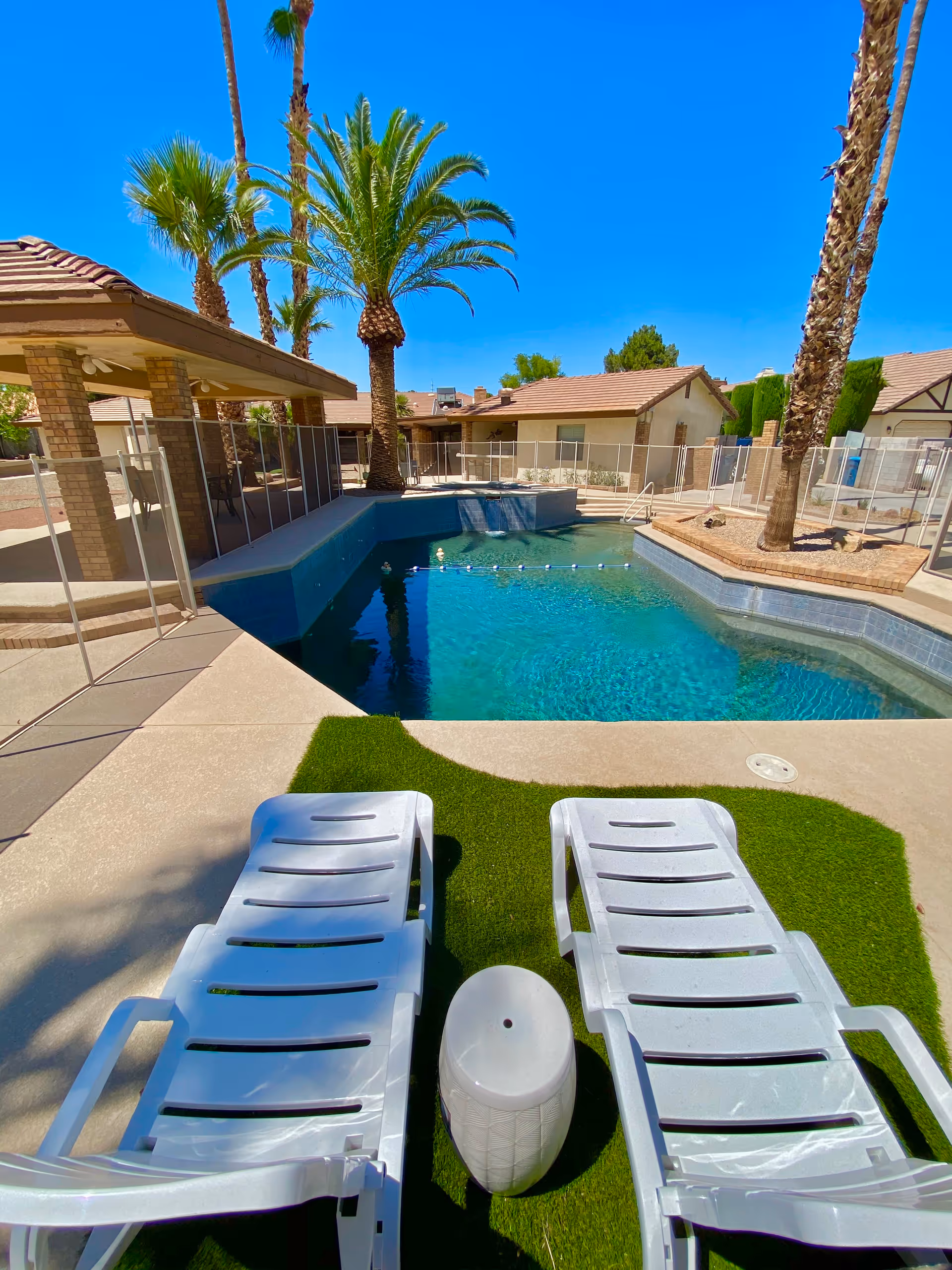 Outdoor swimming pool area with two white lounge chairs on artificial grass, a small white side table between them, palm trees around the pool, and a covered patio with brick pillars in the background under a clear blue sky.