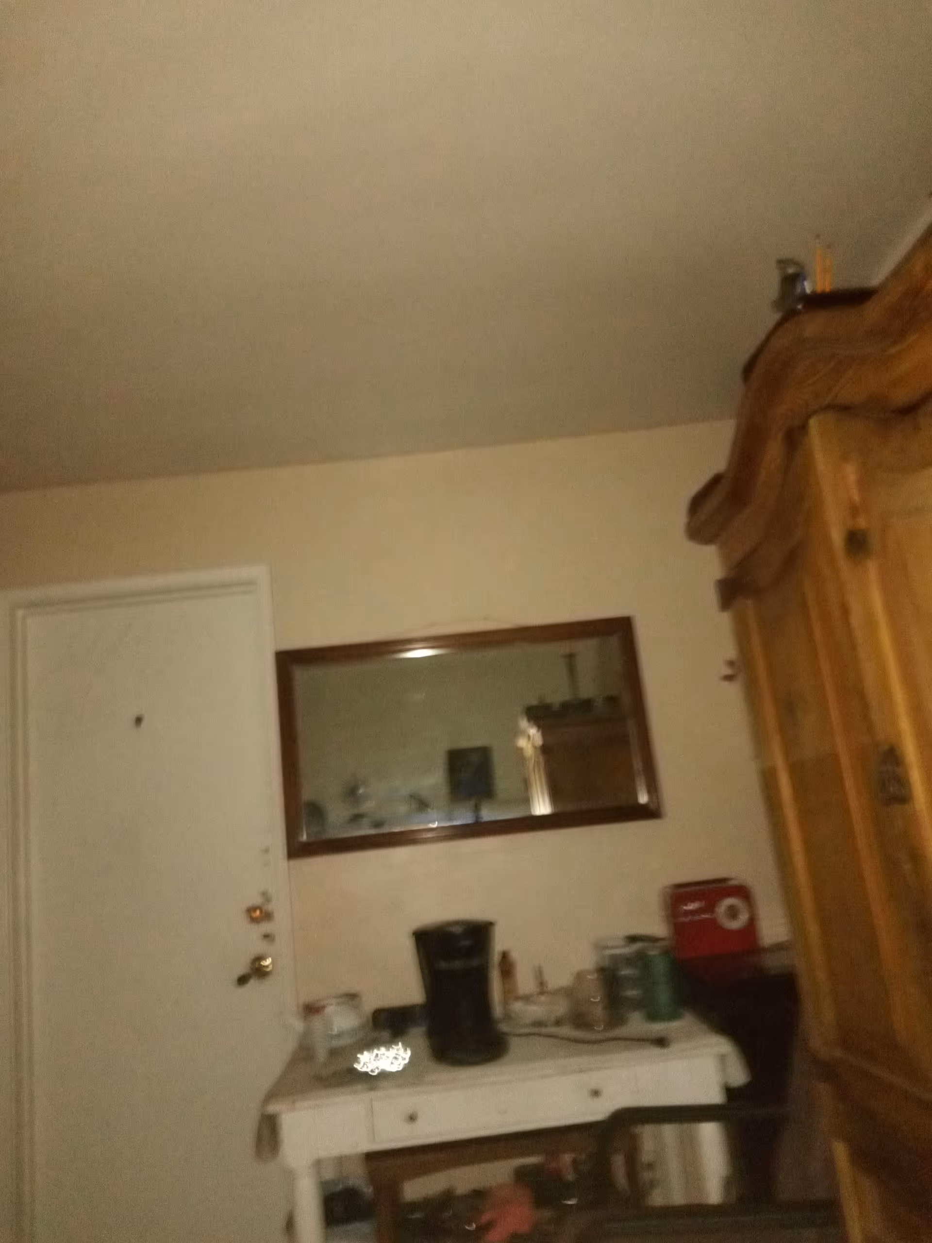 Dimly lit, cluttered entryway with a white dresser, coffee maker, and a large wooden armoire.