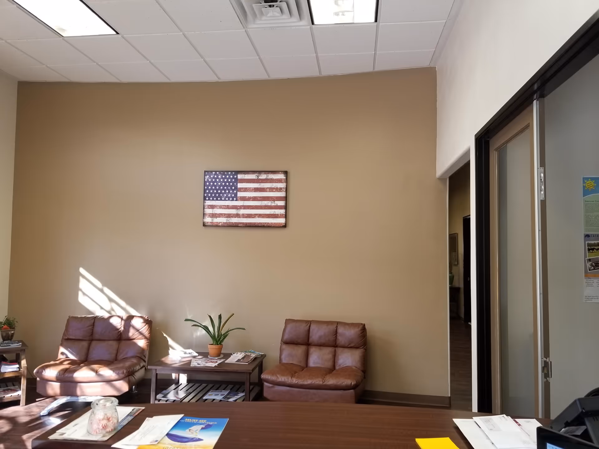 Interior lobby area with beige walls, two brown leather chairs, plant, and American flag artwork.
