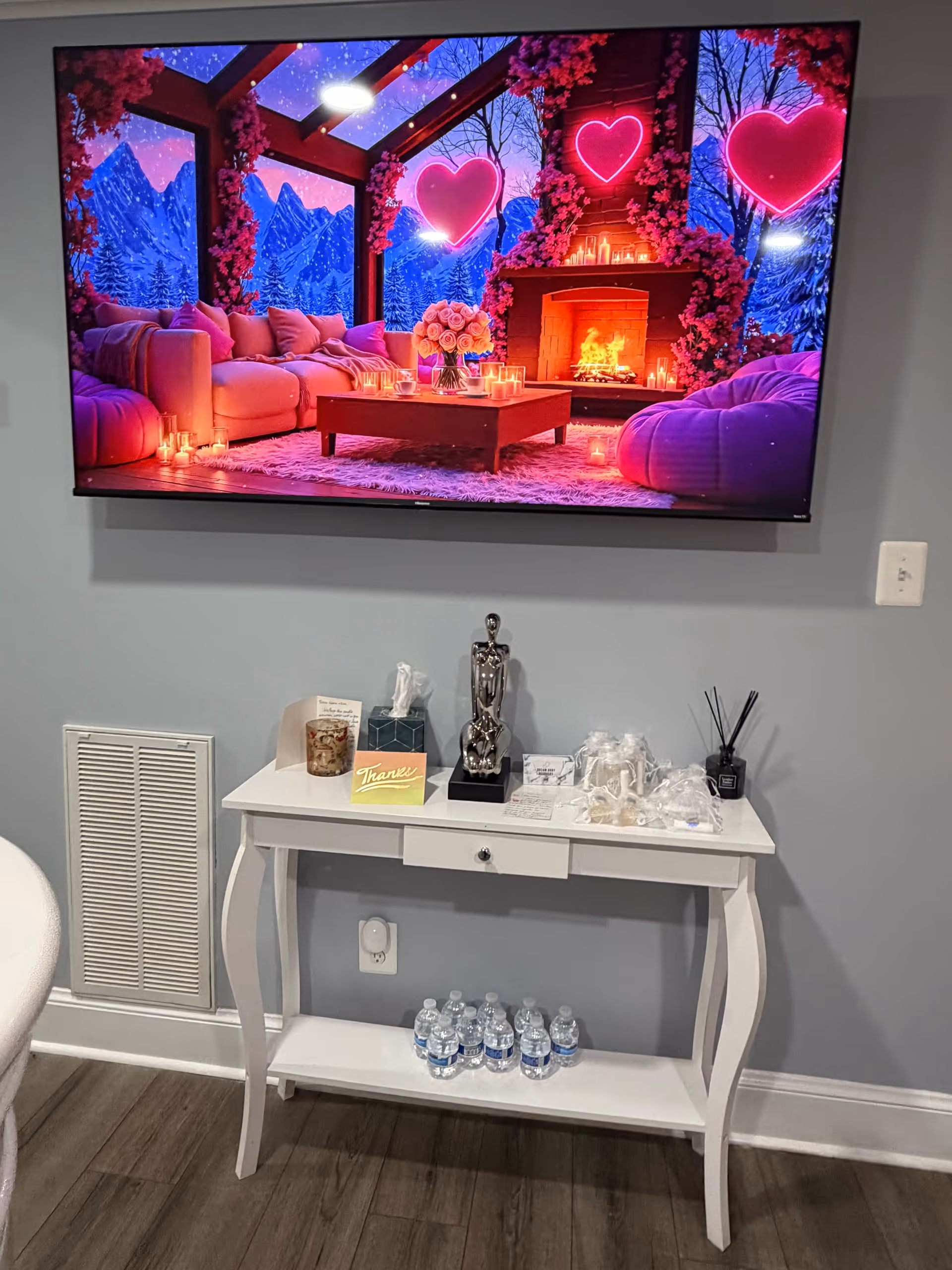 TV displays a cozy living room scene; white console table holds cups, tissues, and water.