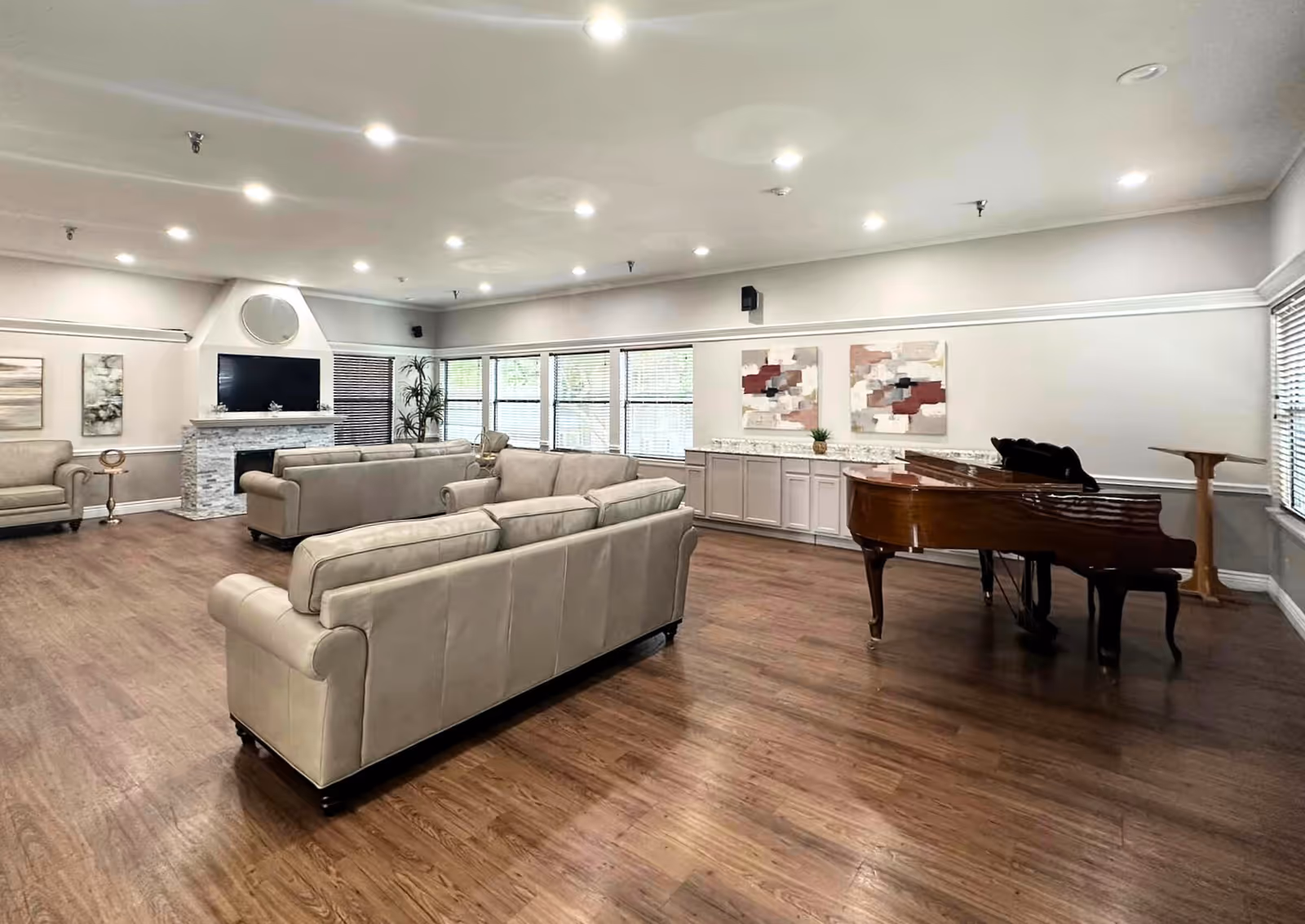 A spacious living room with wooden flooring featuring beige sofas arranged around a white stone fireplace with a mounted TV above it. Large windows with blinds allow natural light to fill the room. There is a grand piano on the right side near a wall with two abstract paintings and a long countertop with cabinets underneath. The ceiling has multiple recessed lights.