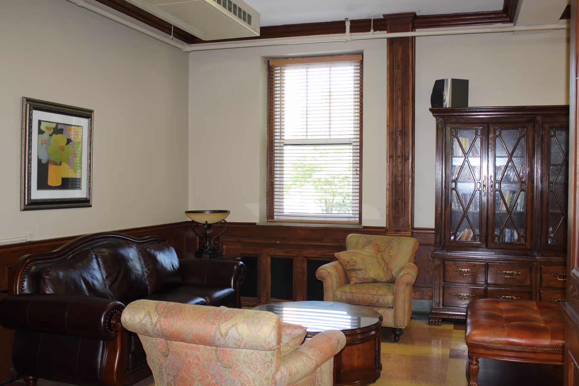 Cozy furnished sitting room with a leather sofa, patterned armchairs, a wooden coffee table, and a glass-front cabinet beneath a window.
