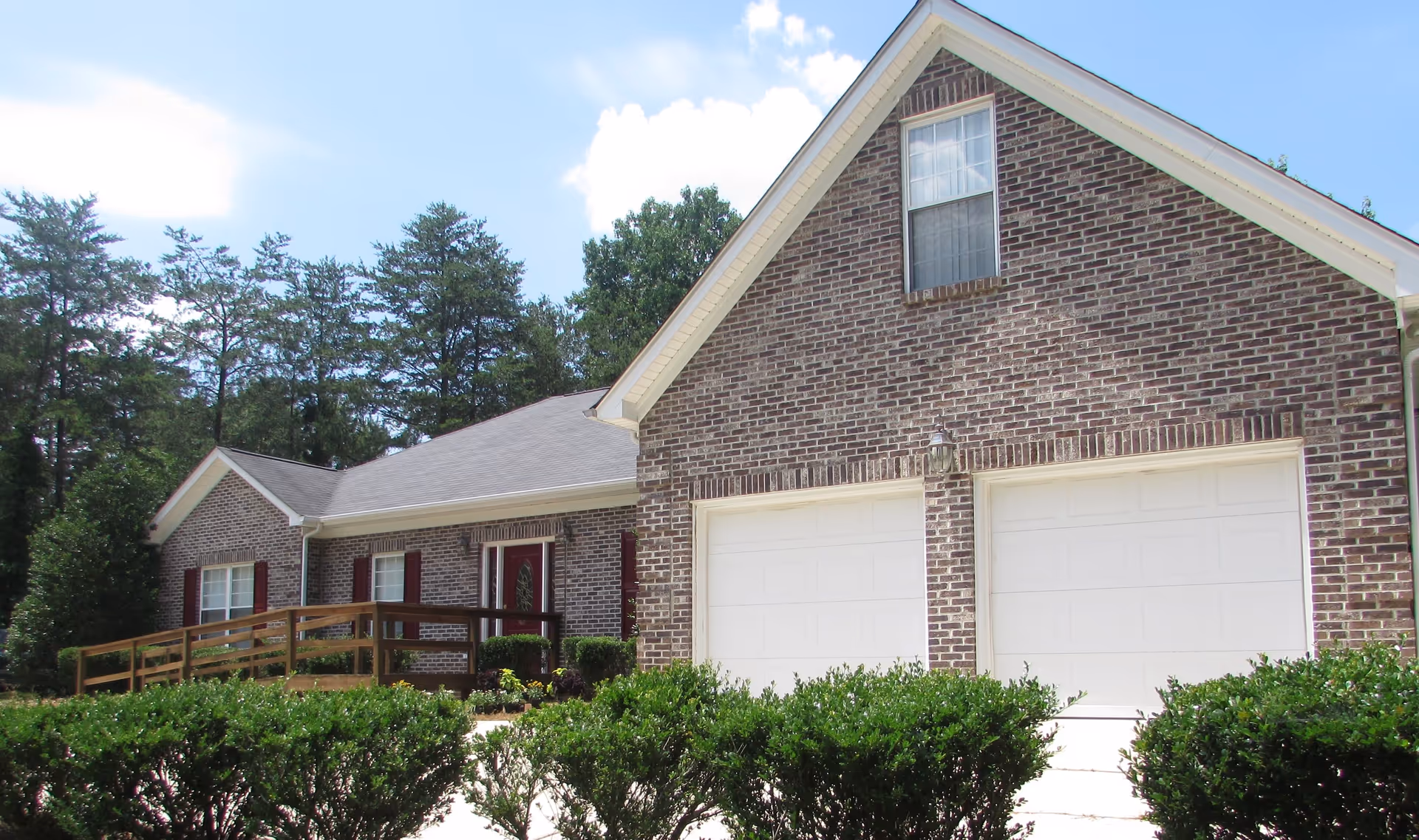Exterior view of a brick building with a double garage door, a front entrance with a wooden ramp, and windows with red shutters. There are green bushes in front and tall trees in the background under a partly cloudy blue sky.