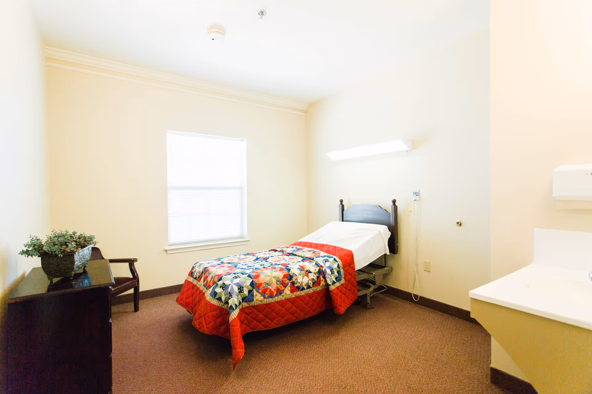A single bed in a small patient bedroom with a quilt, window, dresser, and sink.