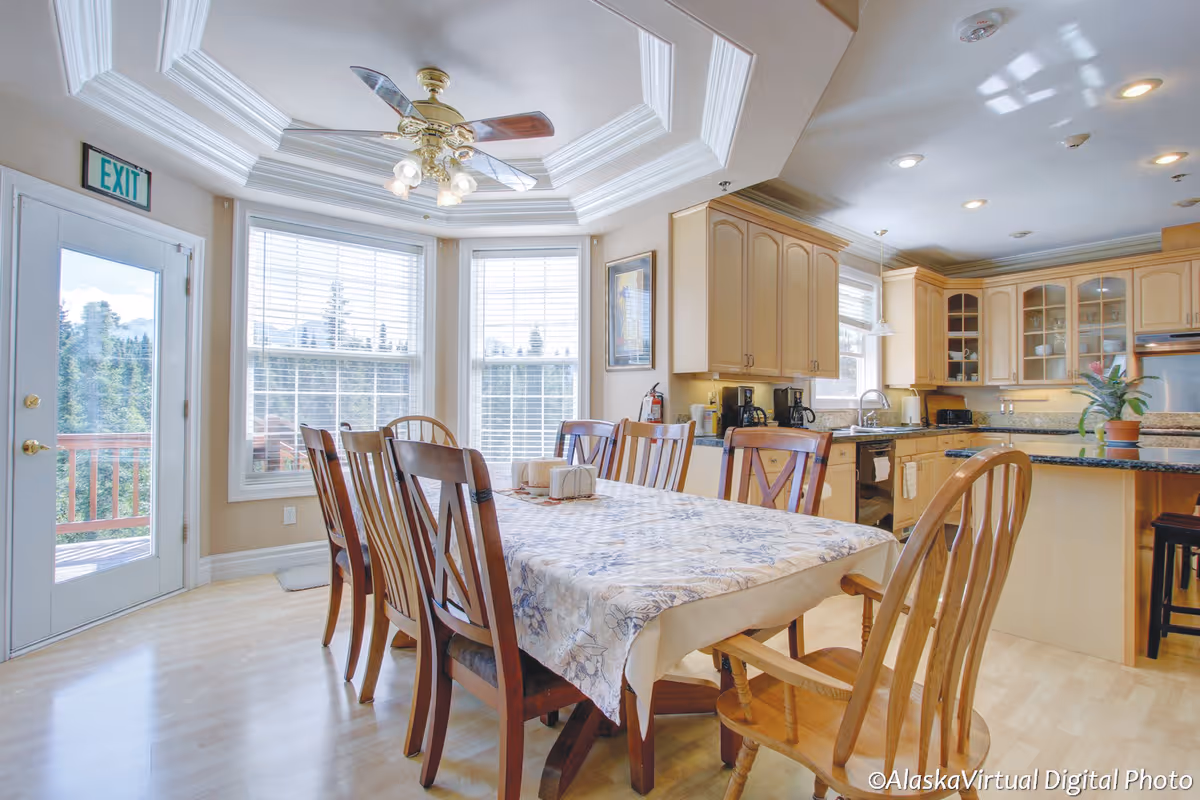 Bright dining area with a long table covered by a floral tablecloth surrounded by wooden chairs. The room features large windows with white blinds, a ceiling fan with lights, and an adjacent kitchen with light wood cabinets and granite countertops. A door leads outside to a wooden deck with a view of trees.