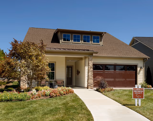 Front exterior view of a single-story house with a brown shingled roof, beige siding, stone accents around the entrance, a two-car garage with dark brown doors, a well-maintained lawn, flower beds, and a concrete walkway leading to the front door. A sign in the yard reads 'Refresh 1903 sq. ft.'