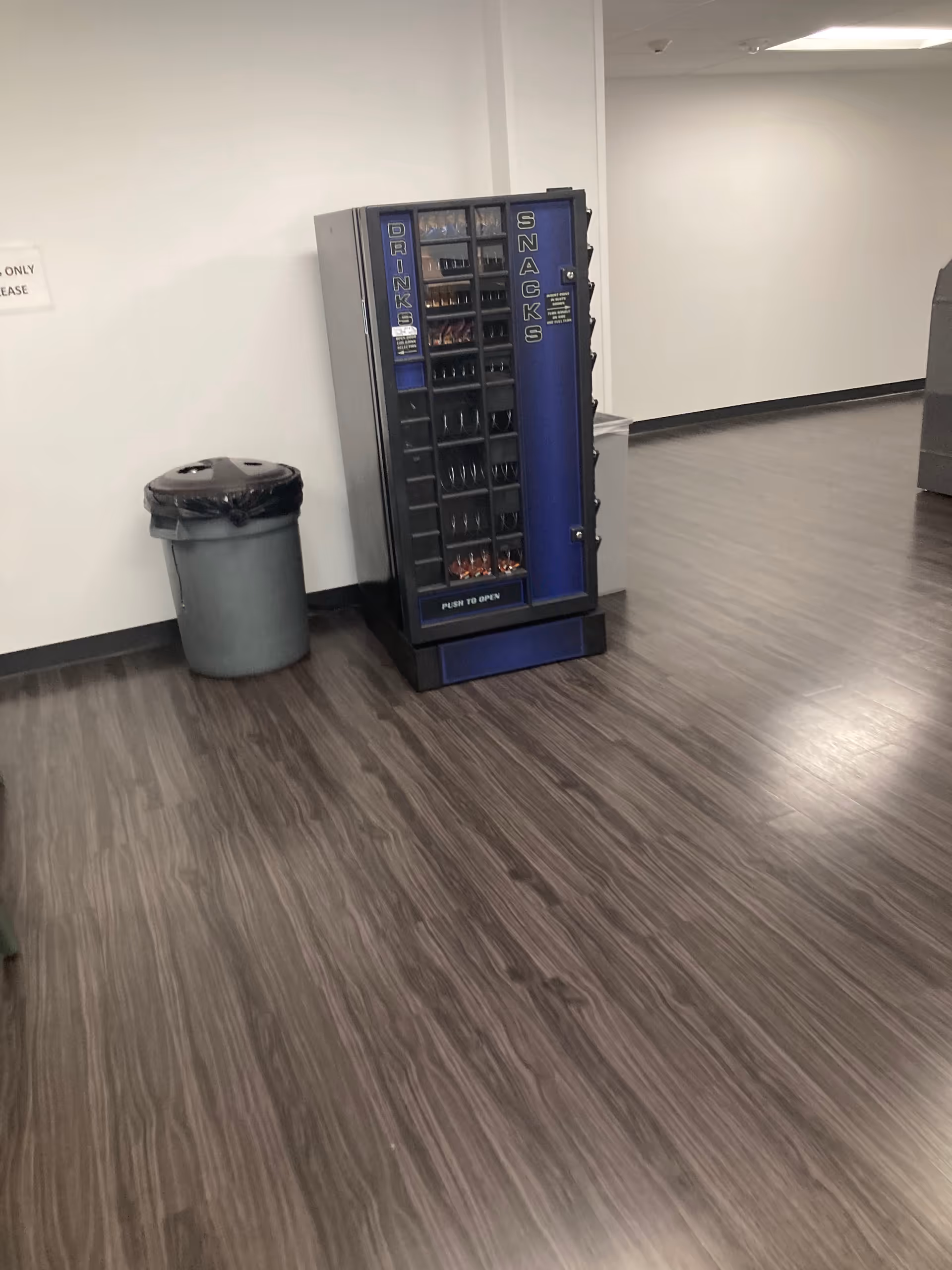 Blue vending machine beside a trash can in a quiet hallway of a senior facility.