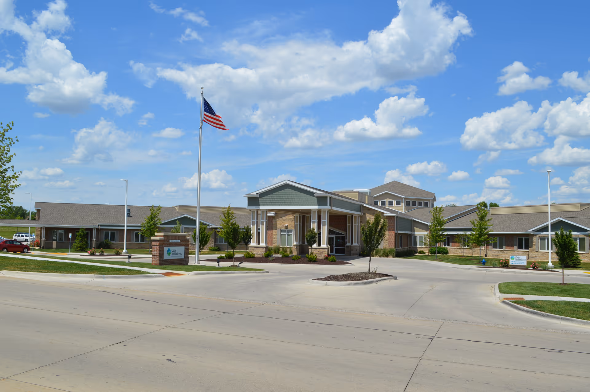 Entrance and front exterior of a single-story senior care building with an American flag and a circular driveway under a blue sky.