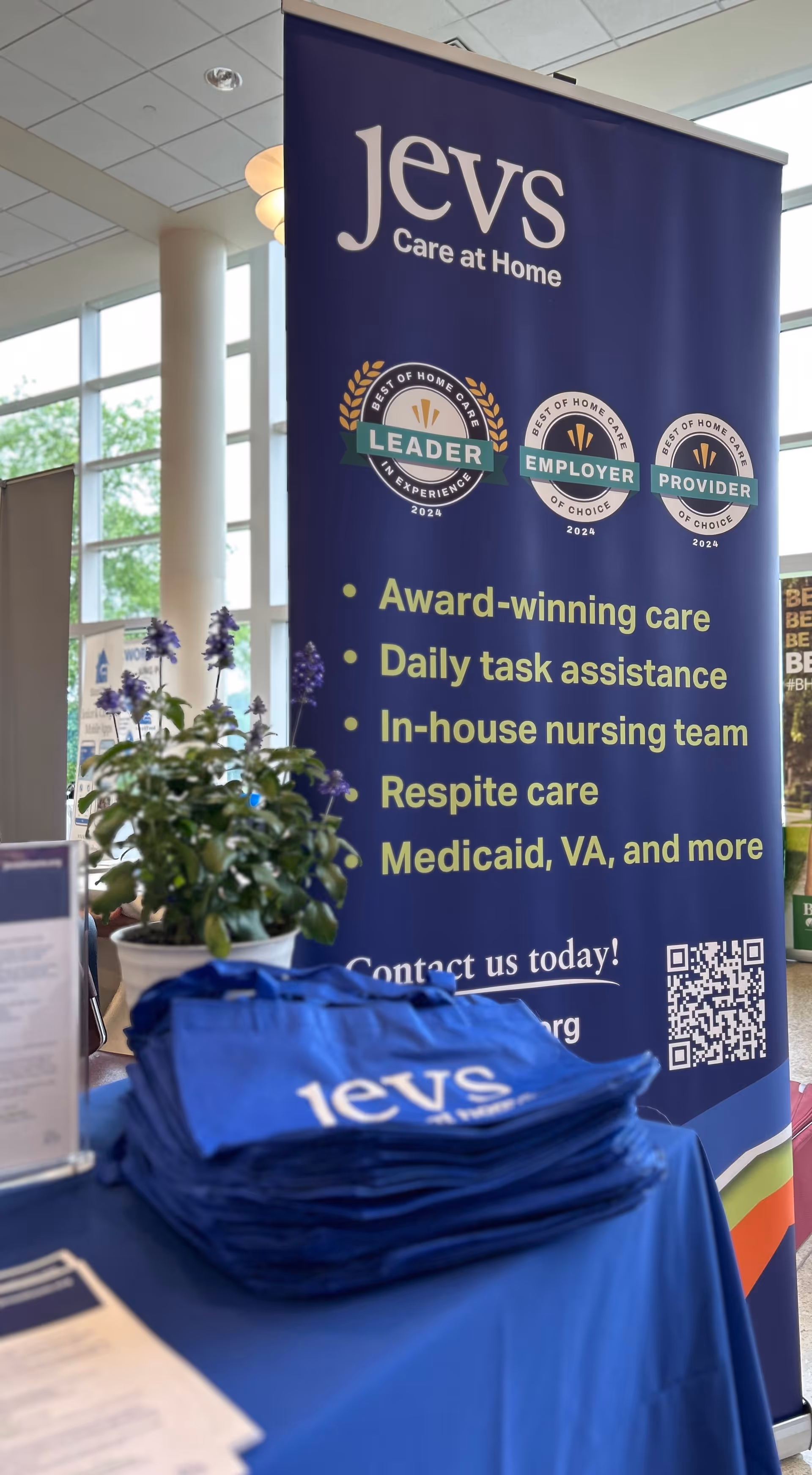 Indoor promotional banner for JEVS Care at Home with blue table, plants, and tote bags.