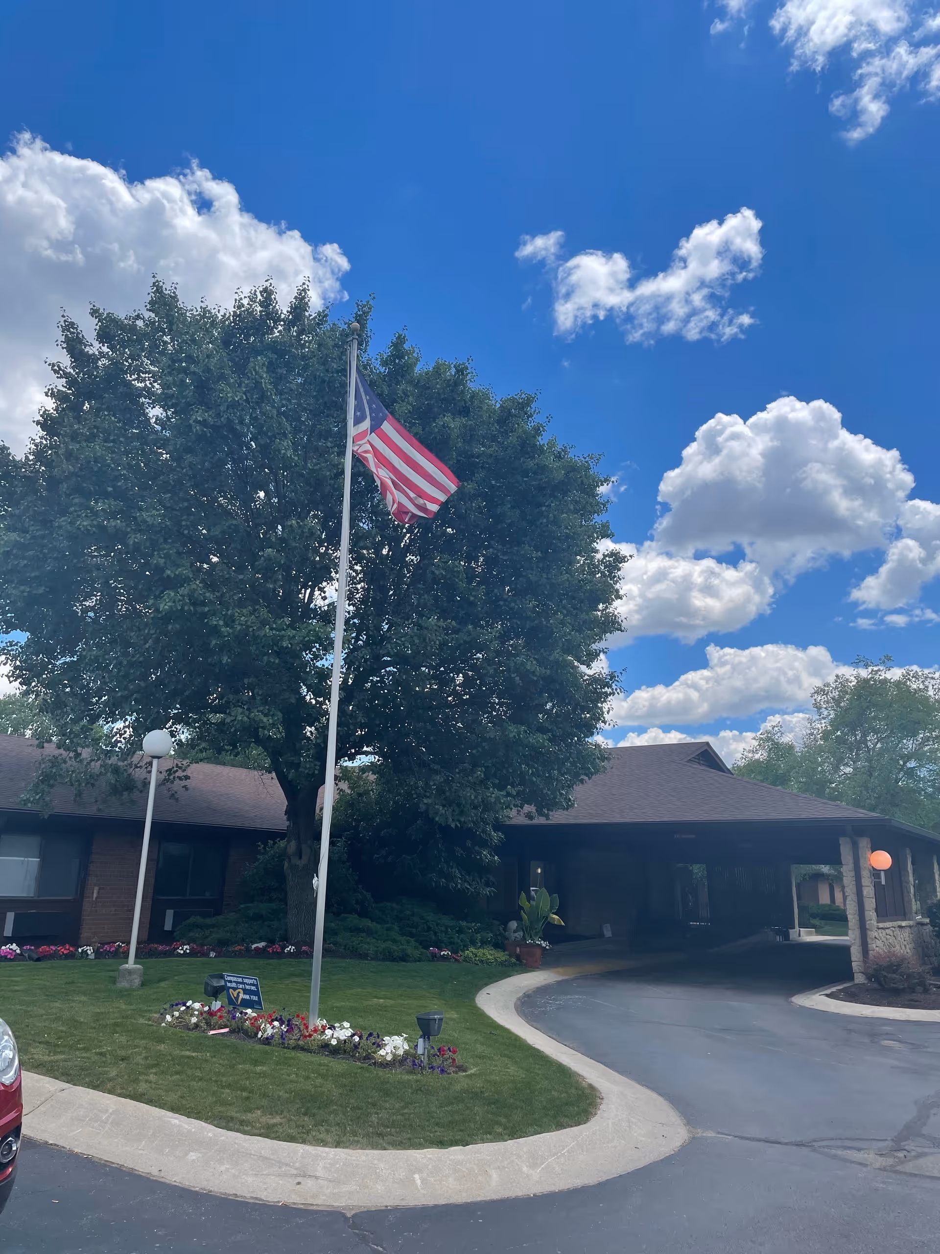 Front entrance of a senior living facility with an American flag on a pole, circular driveway, and canopy under a blue sky.