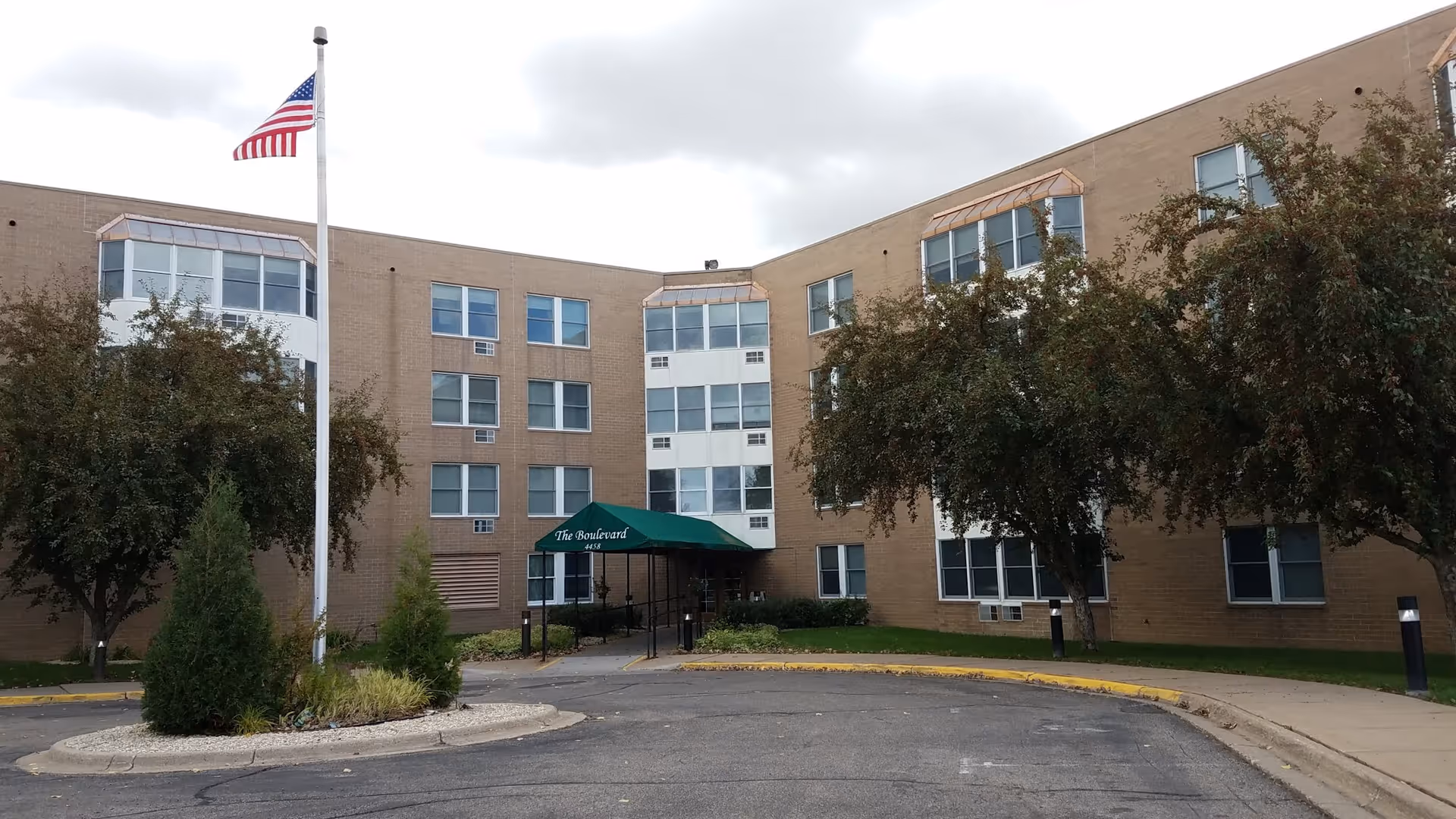 Exterior view of a three-story senior living facility building with a beige brick facade. There is a green awning over the entrance that reads 'The Boulevard'. An American flag is flying on a flagpole in front of the building, and there are trees and shrubs around the entrance area.