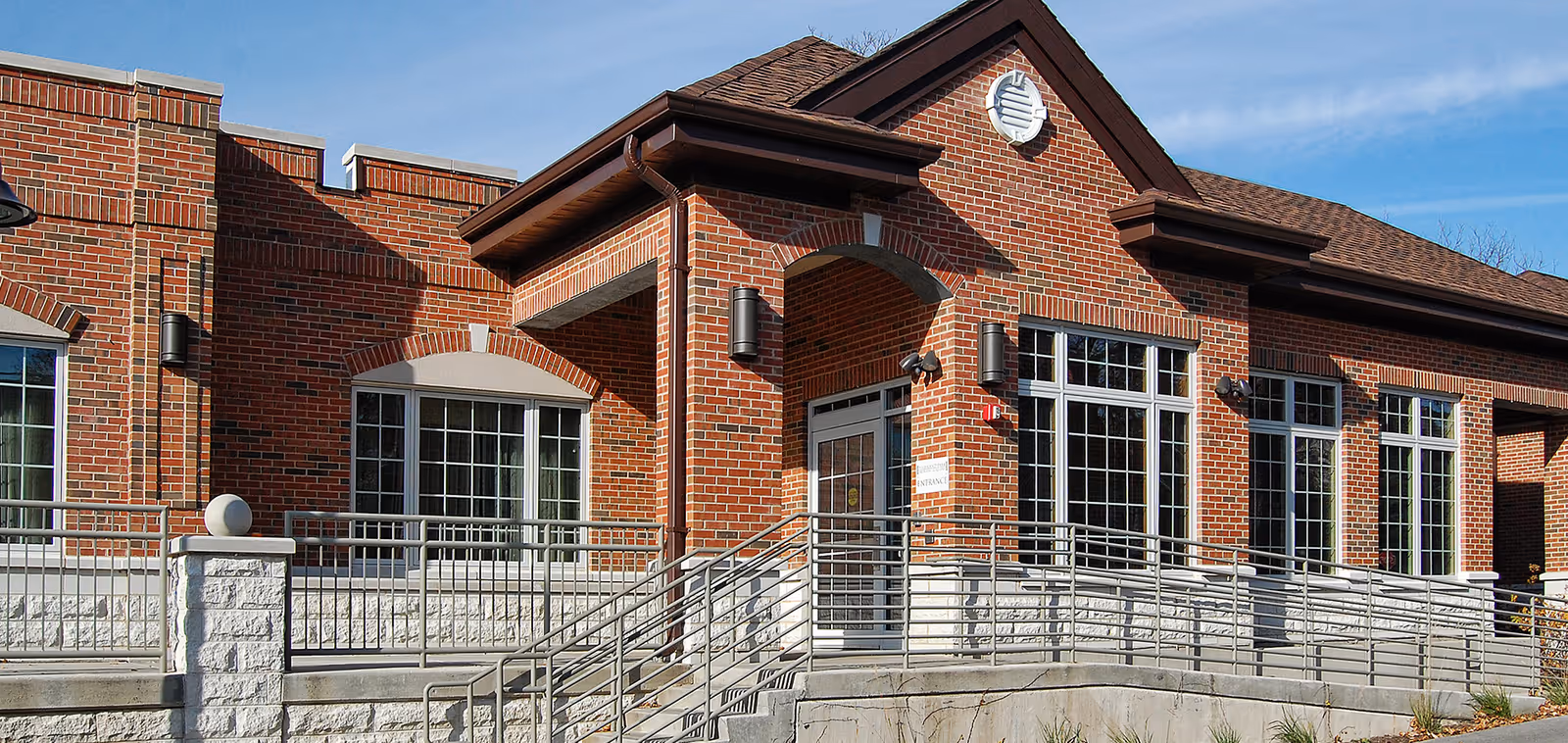 Exterior view of a brick building with large windows and a ramp leading to a glass door entrance under a covered porch, set against a clear blue sky.