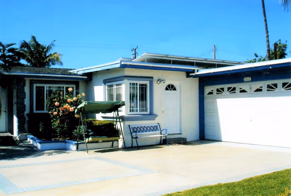 Single-story light-colored house front with a two-car garage, driveway, porch bench, swing, and flowering bushes under a clear blue sky.