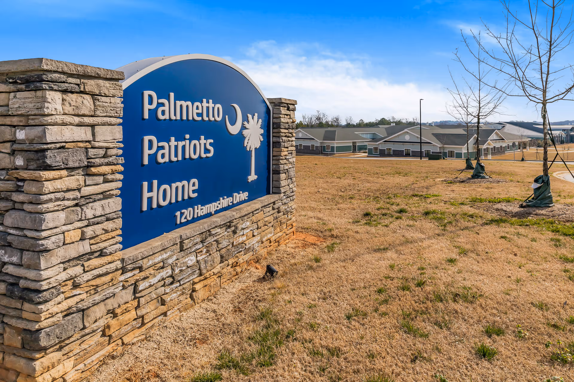 Blue Palmetto Patriots Home sign with stone pillars and campus buildings in background.